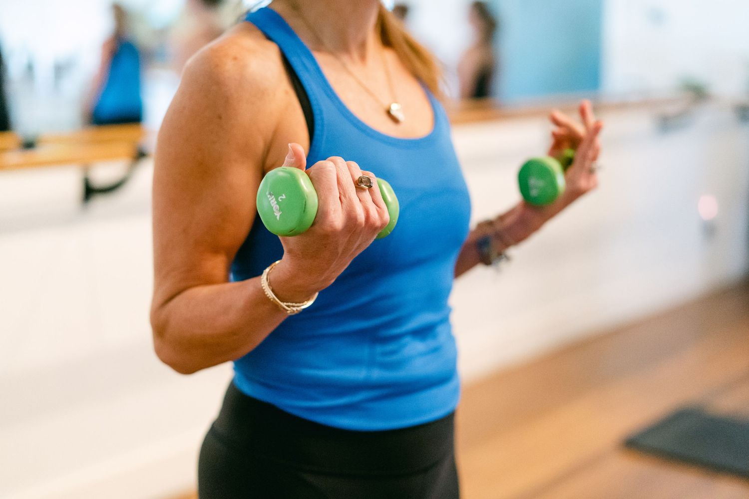 Woman in blue tank top doing bicep curls with green dumbbells during fitness class.