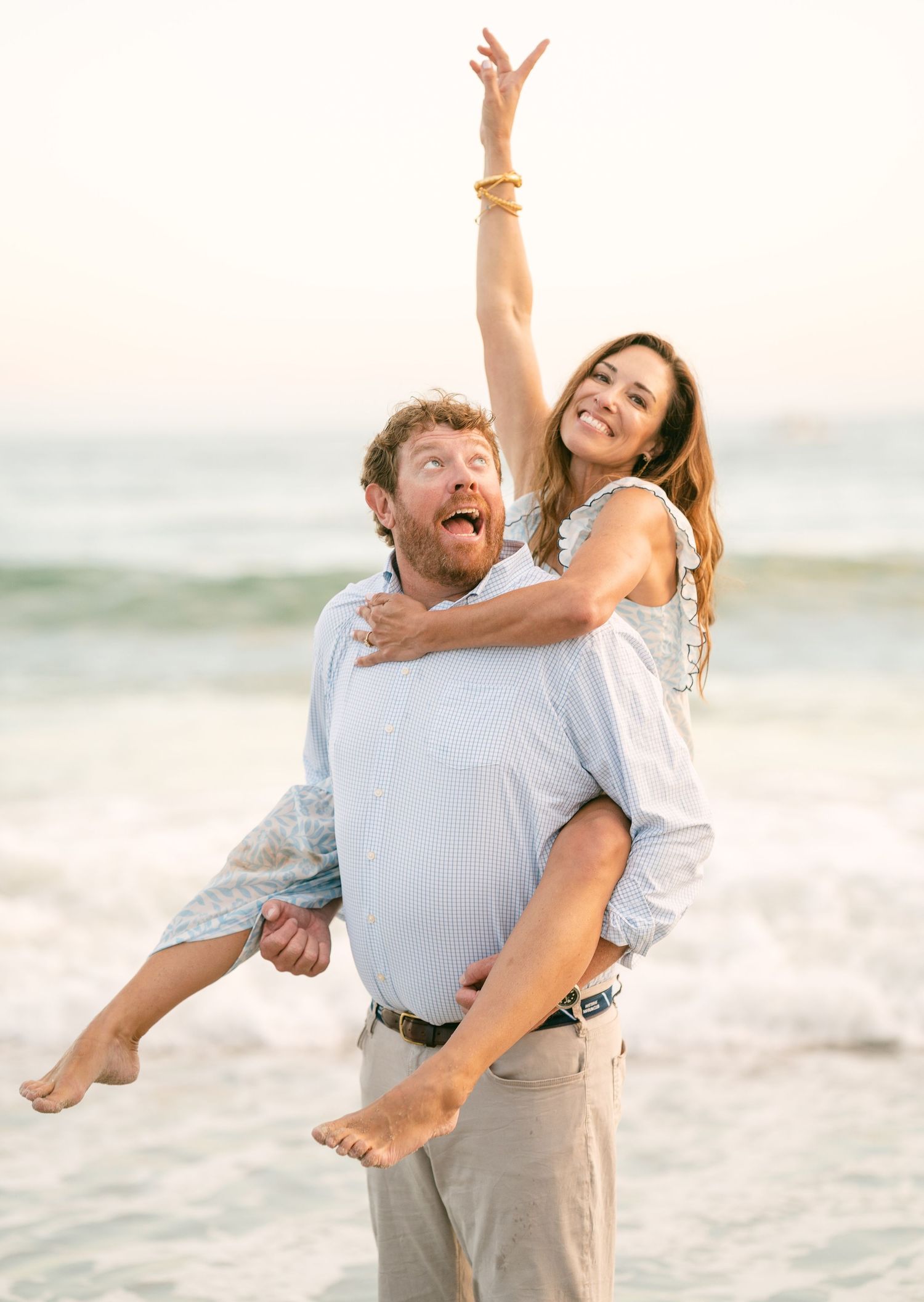 Joyful couple at beach, man giving woman piggyback ride, both laughing, woman raising arm in celebration.