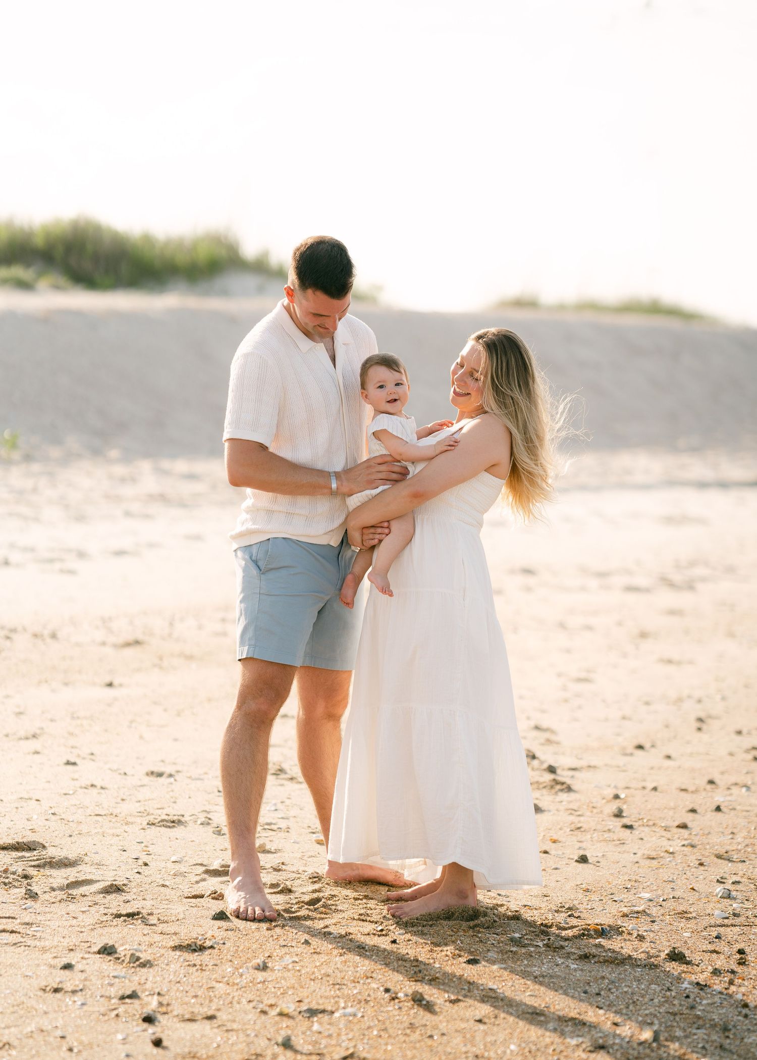 Young couple in white outfits holding smiling baby on sandy beach at golden hour sunset.