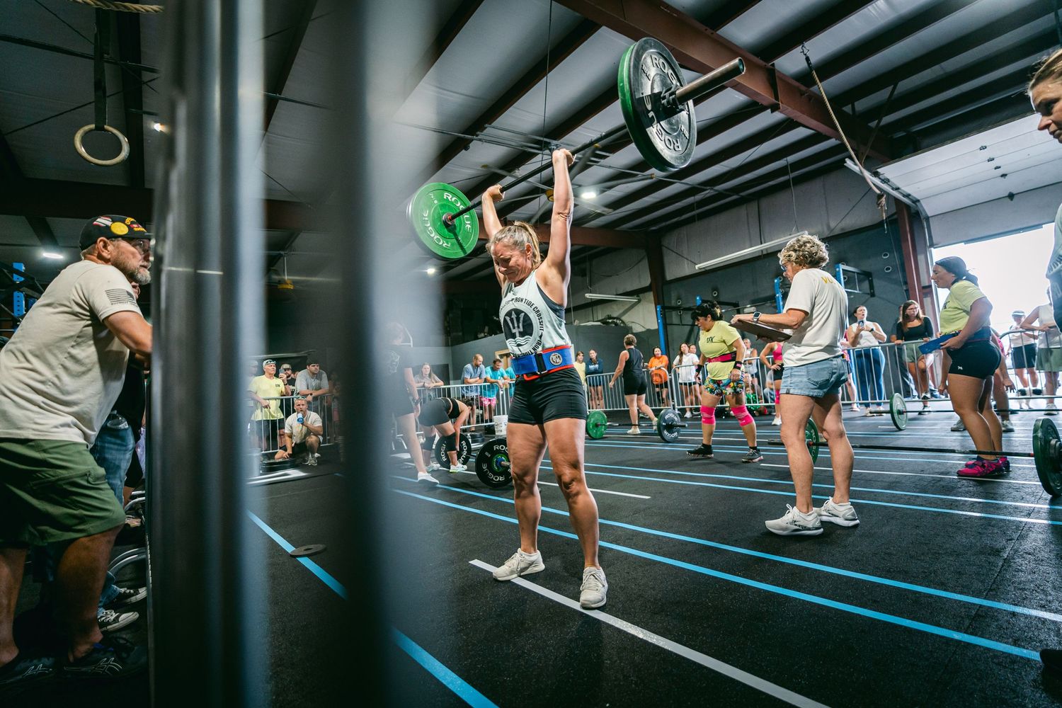 Athlete lifting a barbell overhead during a CrossFit competition inside a gym with spectators.