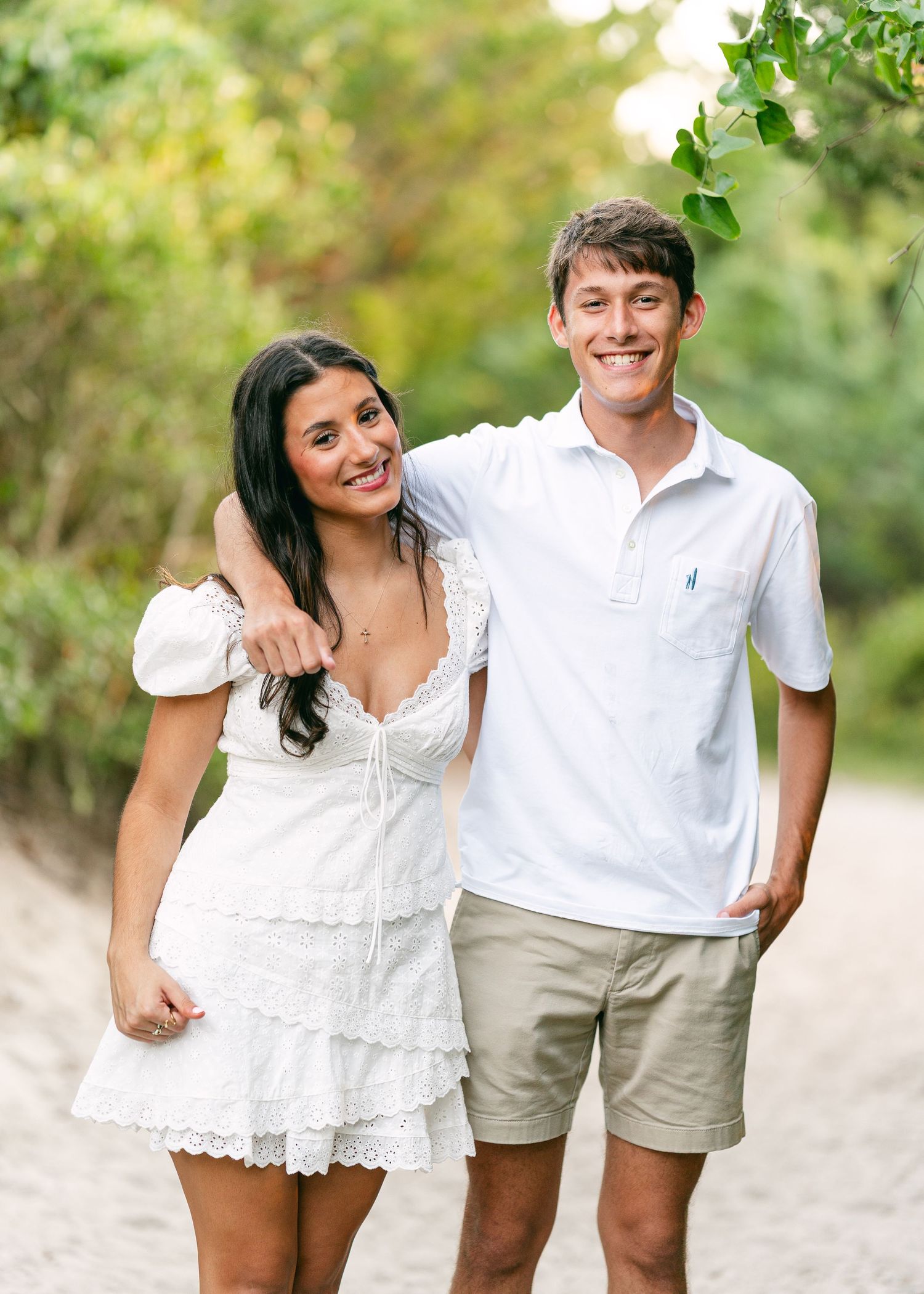Smiling couple in white outfits walking on sandy tropical beach path.