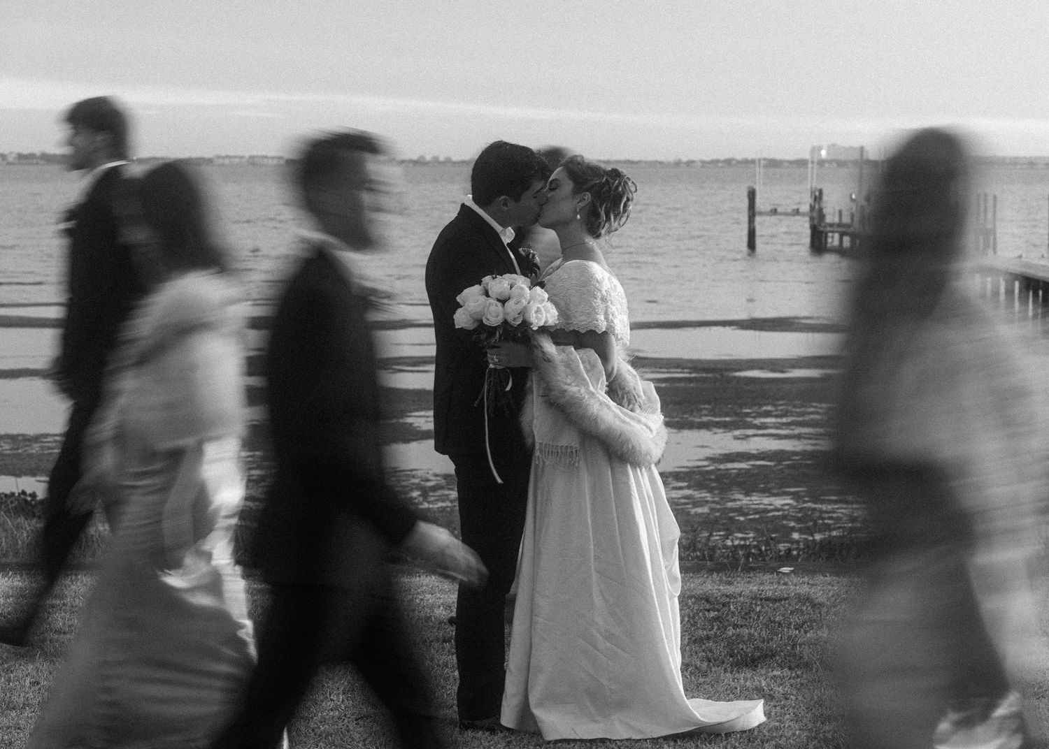 Black and white photo of bride and groom kissing waterside as wedding guests walk past.