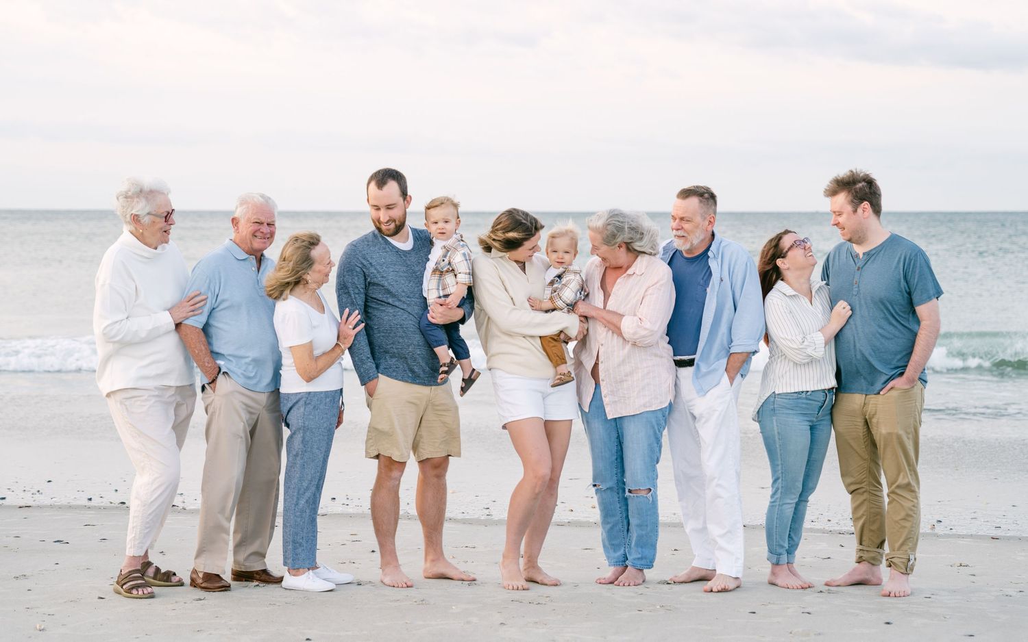 Multigenerational family gathered on beach, interacting warmly, holding babies, dressed in coordinated coastal outfits.