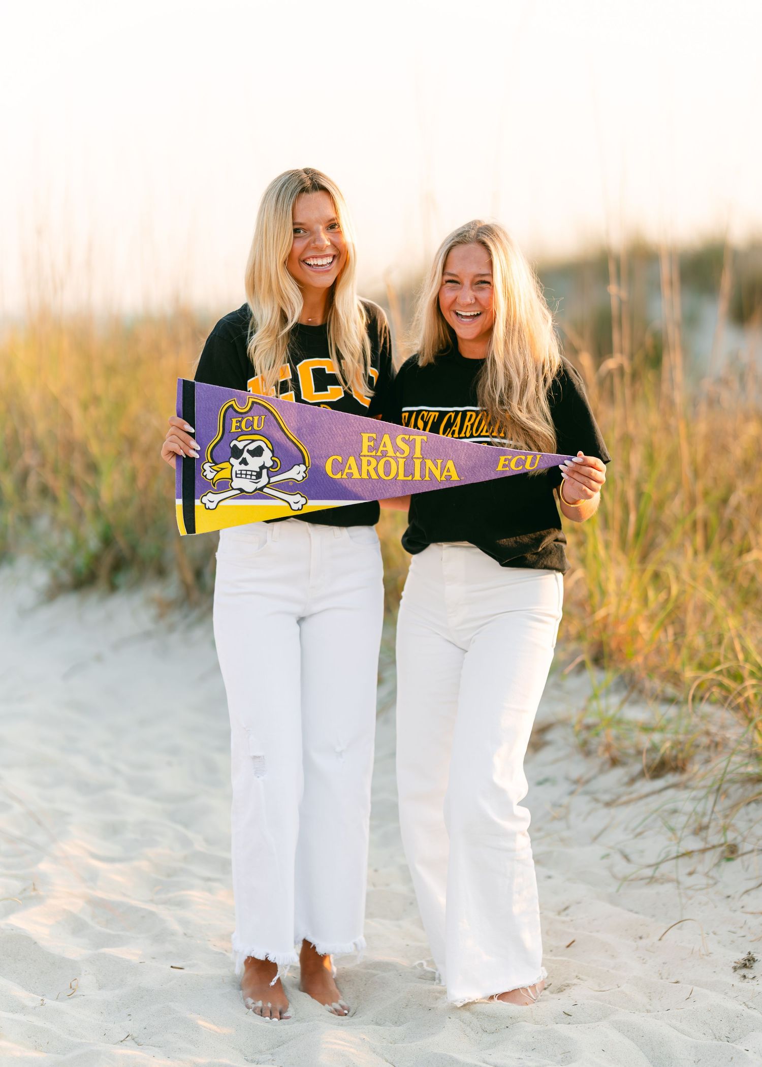 Two smiling blonde women holding ECU East Carolina University pennants on beach at sunset.