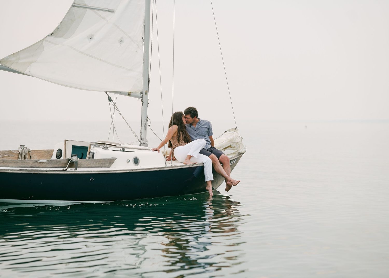 Romantic couple sitting on the bow of a sailboat with feet dangling over calm water.