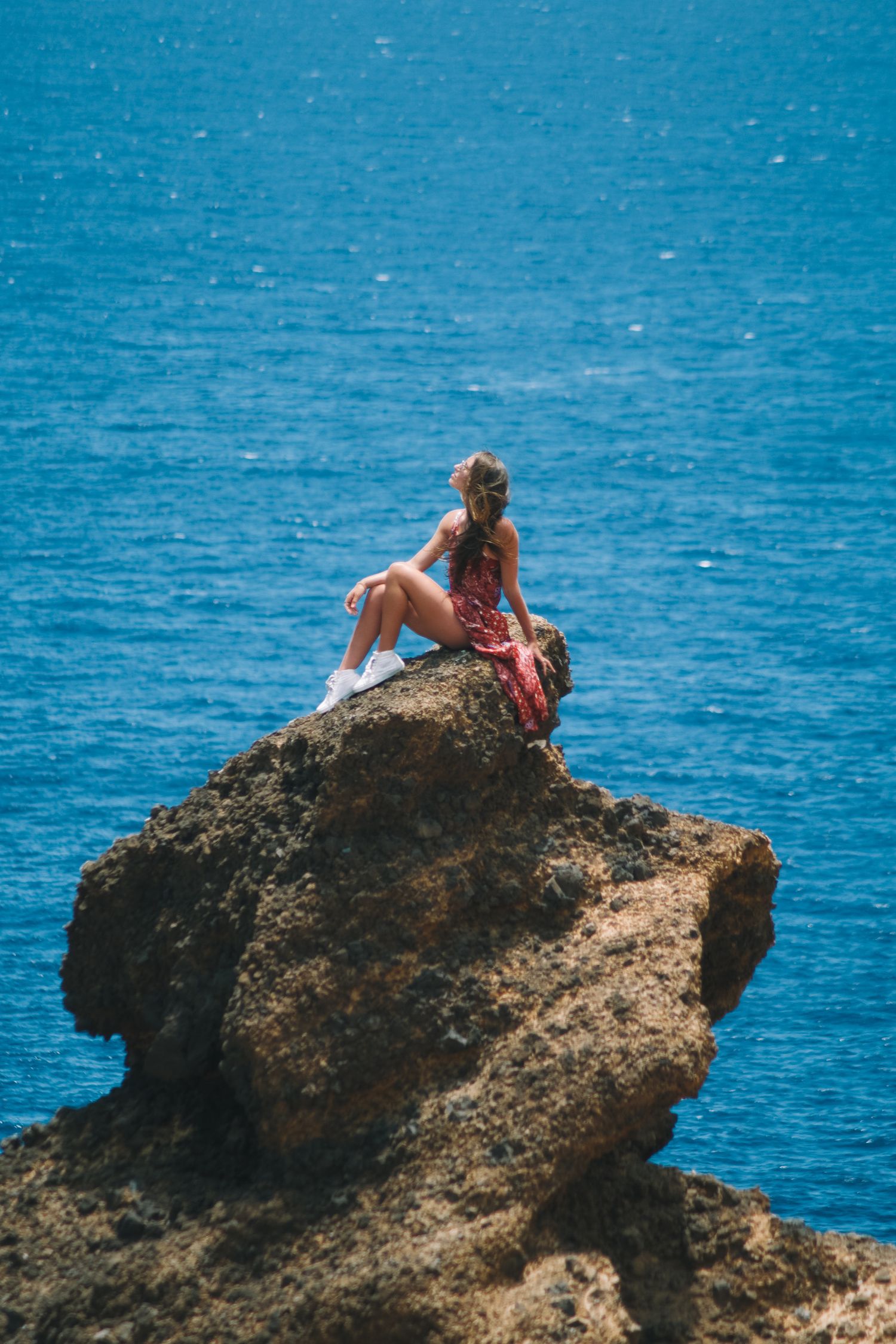 Woman in red floral dress sitting on a rocky cliff overlooking a vivid blue ocean on a sunny day.
