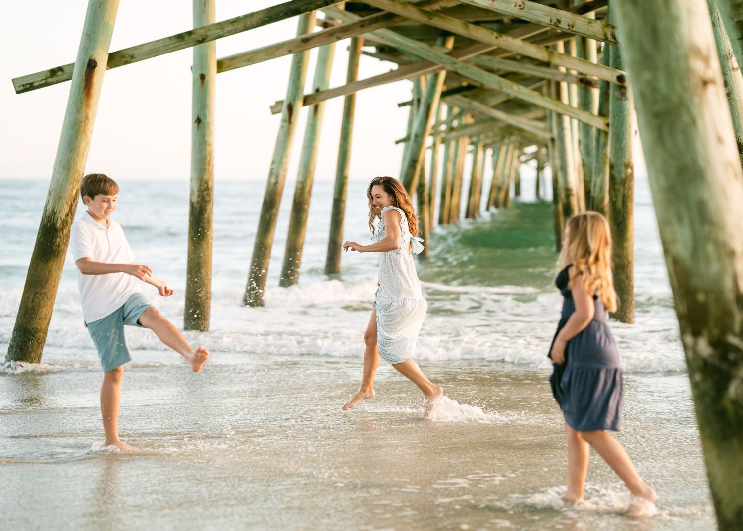 Mother and children playing in ocean waves under beach pier.