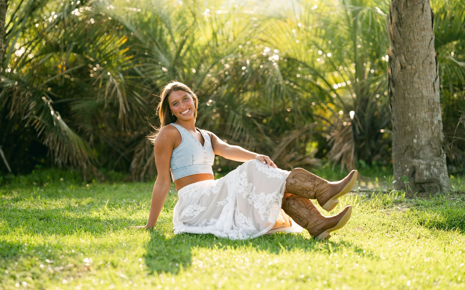 A smiling young woman in a white skirt and blue crop top sits on green grass near palm trees.