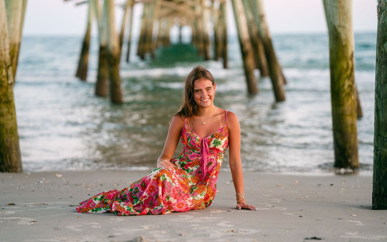 Smiling young woman in colorful floral maxi dress sitting on beach near wooden pier.