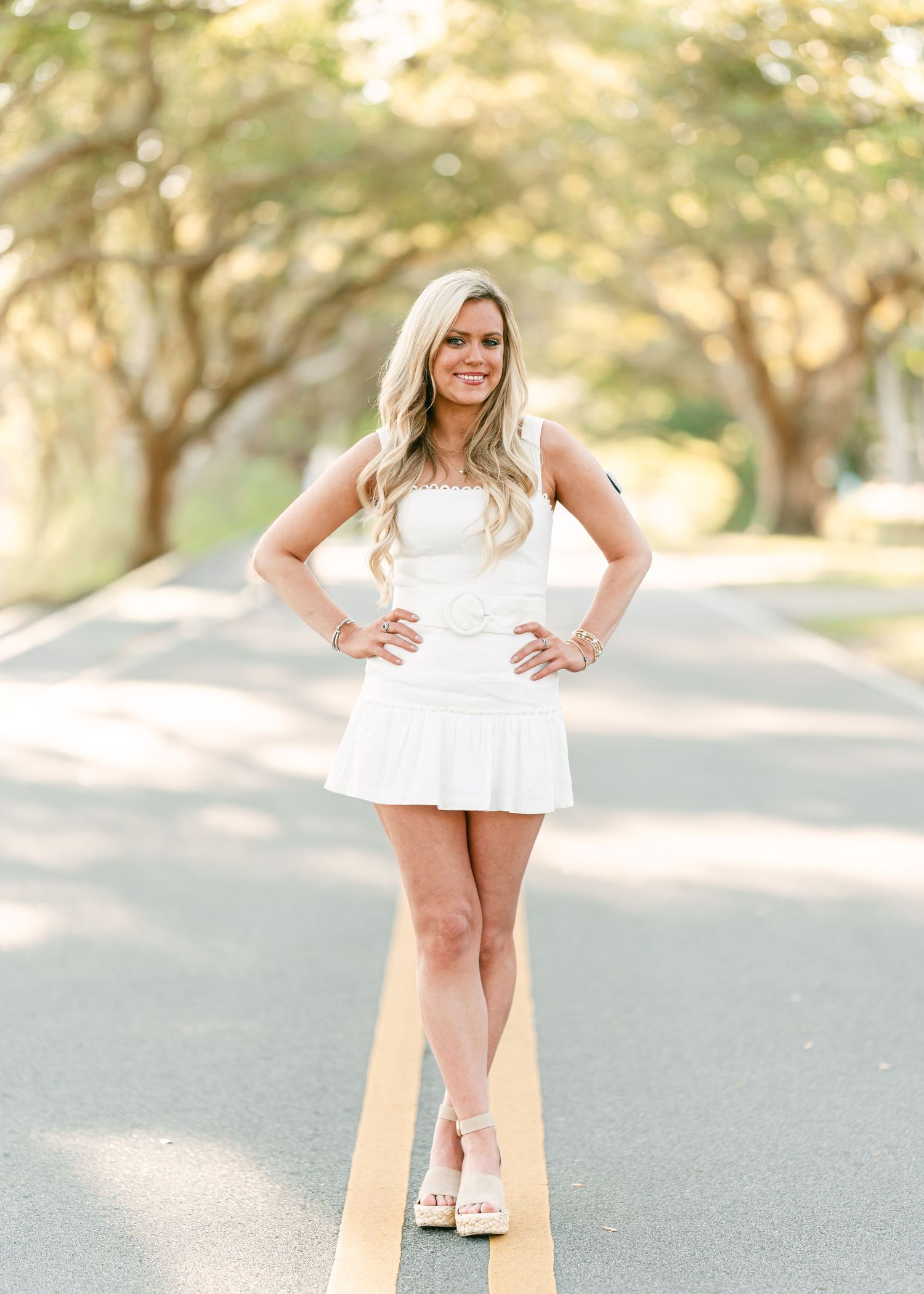 Blonde woman in white dress poses confidently on a tree-lined road center stripe.