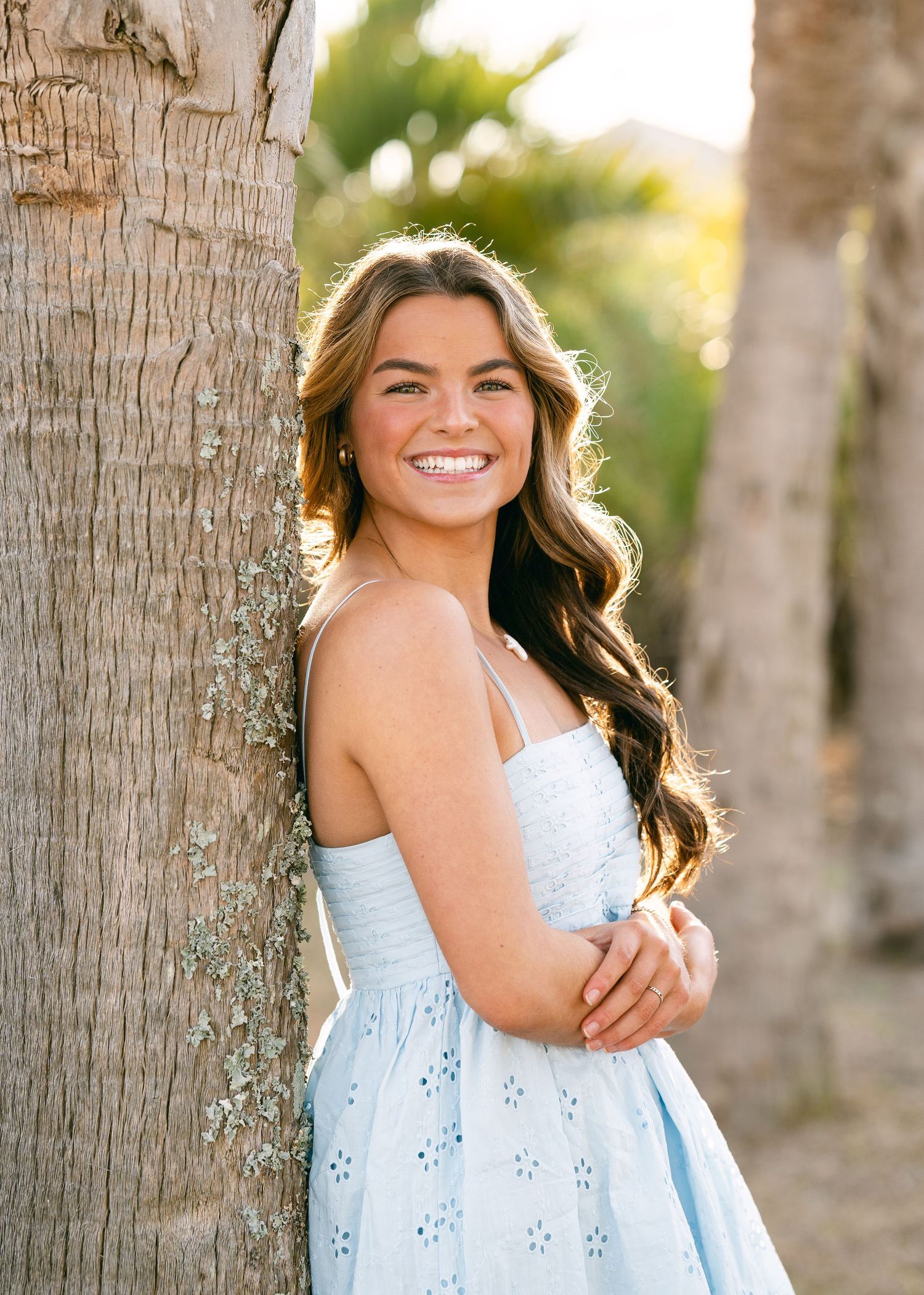 Teenage girl in light blue dress leaning against palm tree smiling for senior portrait.