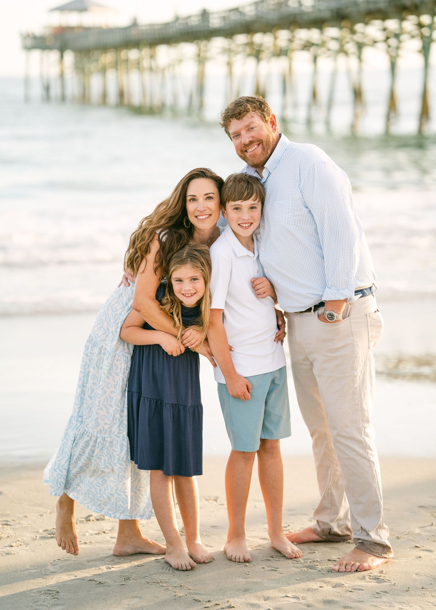 Smiling family of four embracing on beach near wooden pier.