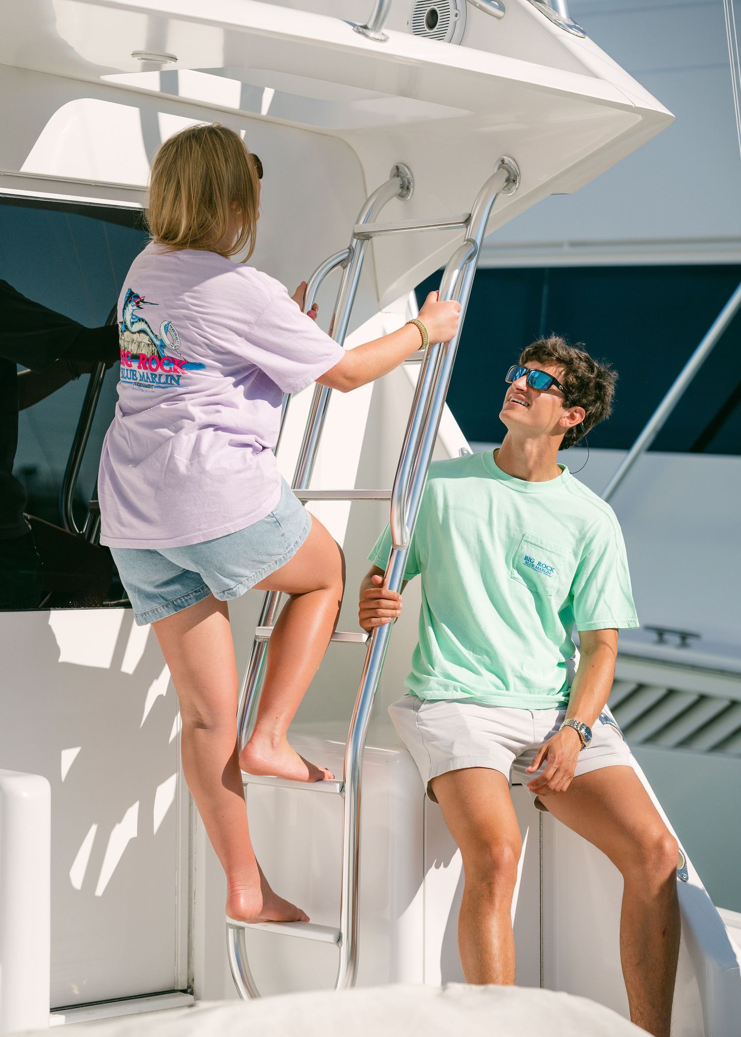 Two young people on a boat, one climbing a ladder in a lavender Big Rock t-shirt.