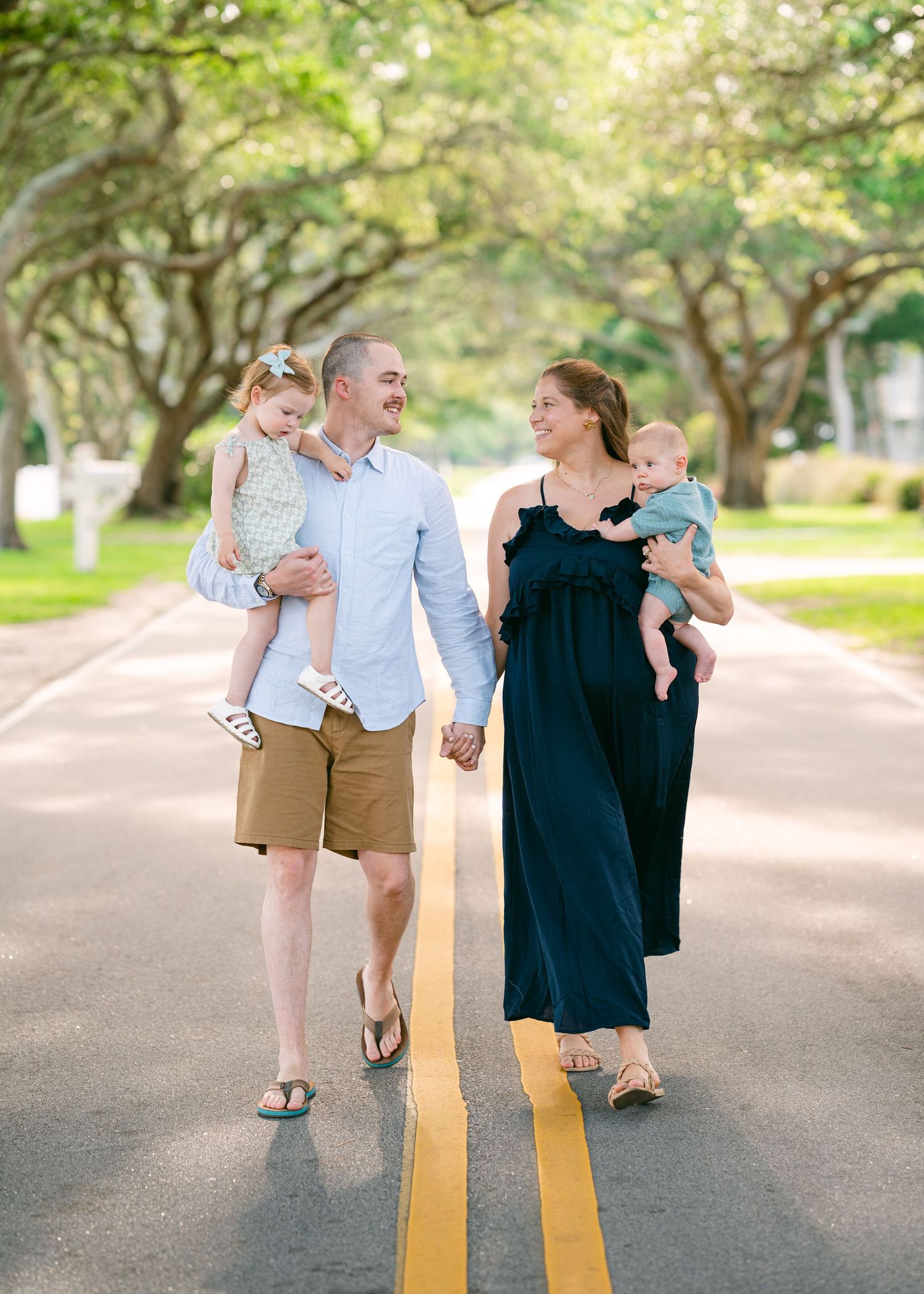 Happy family of four walking hand-in-hand down a tree-lined road, parents carrying young children.