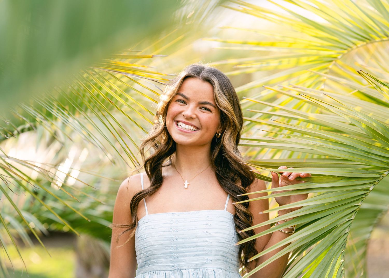 Happy young woman in a light blue dress smiling among lush green tropical palm leaves.