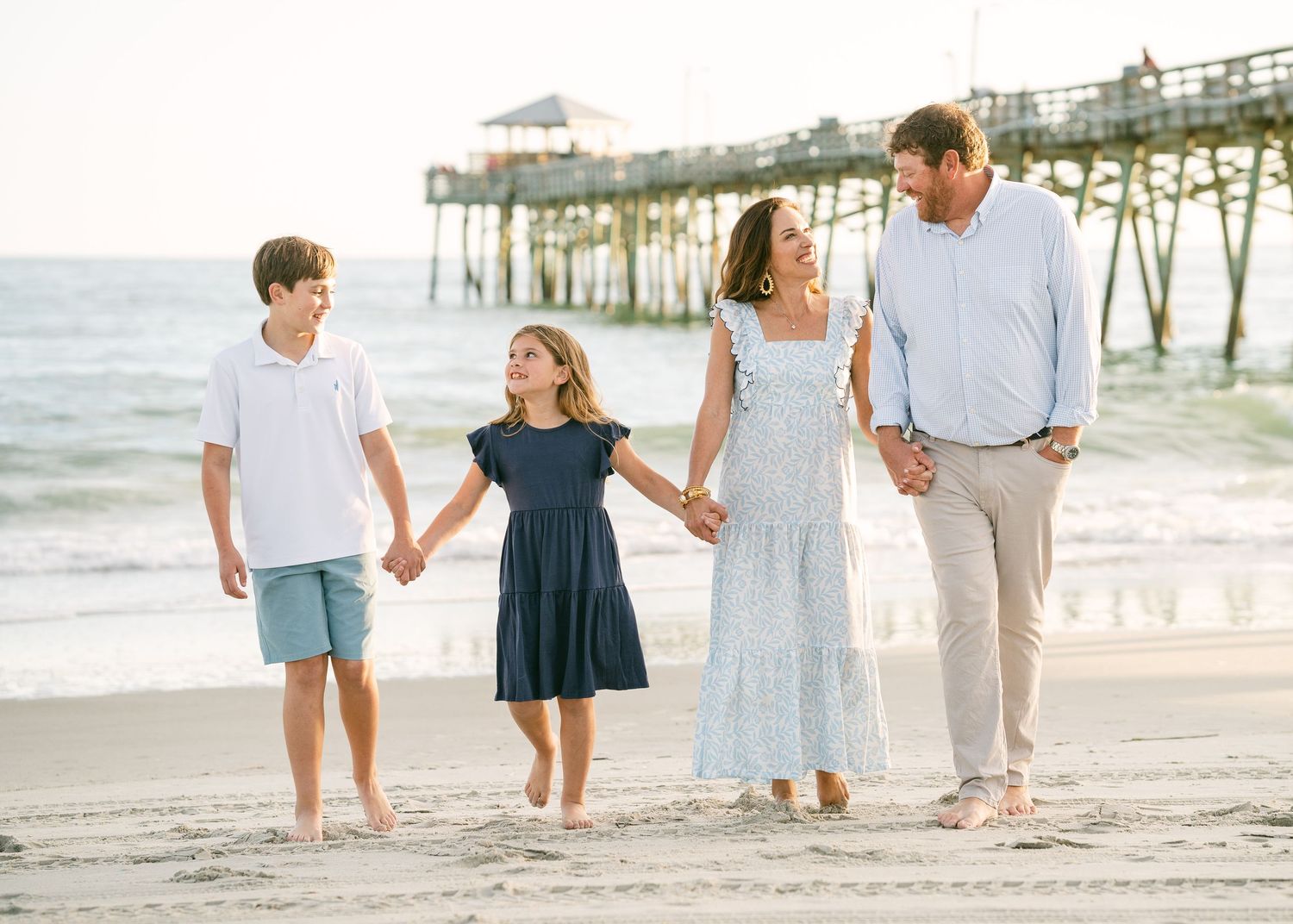 Family of four holding hands walking on beach near pier at sunset.