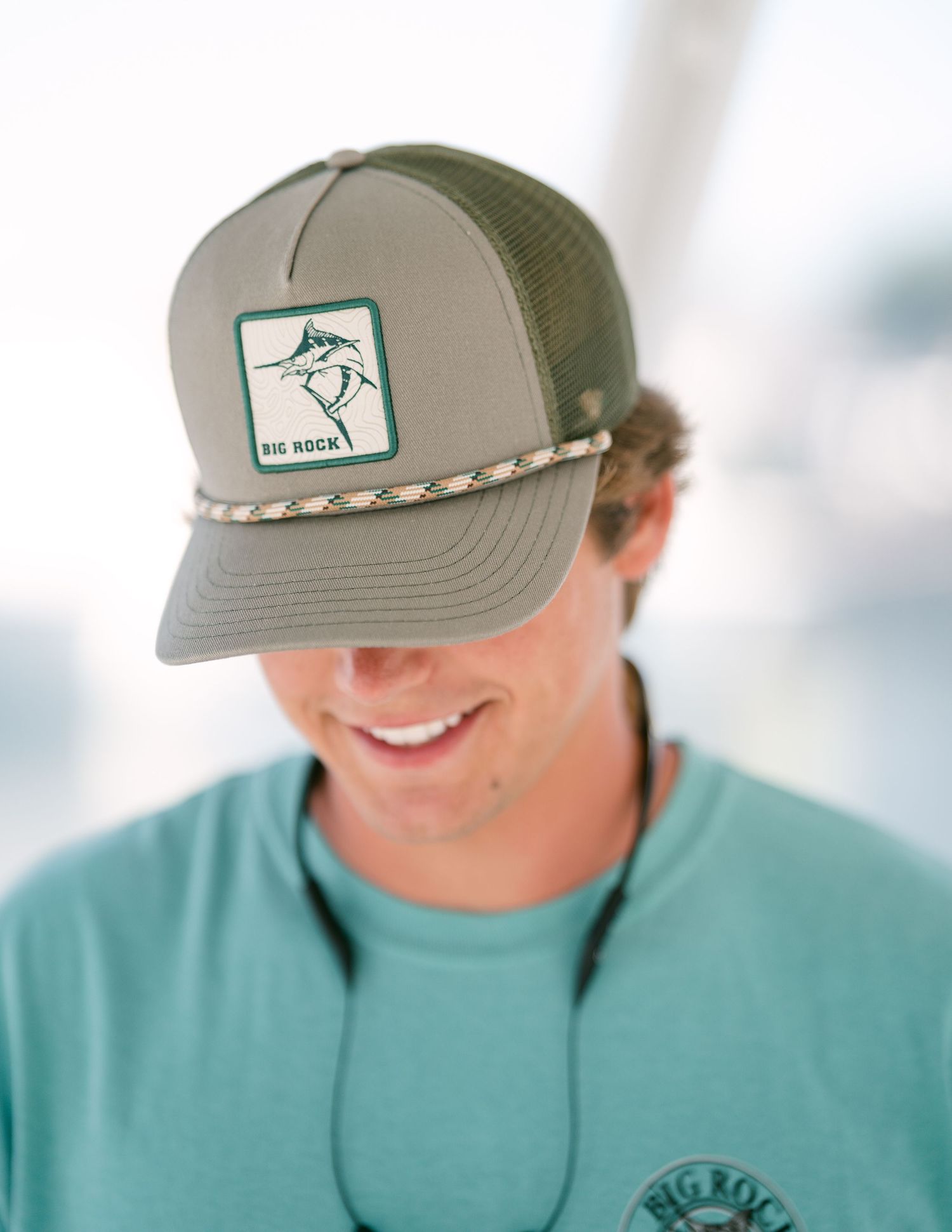 Smiling man wearing an olive green Big Rock trucker hat with marlin logo and teal shirt outdoors.