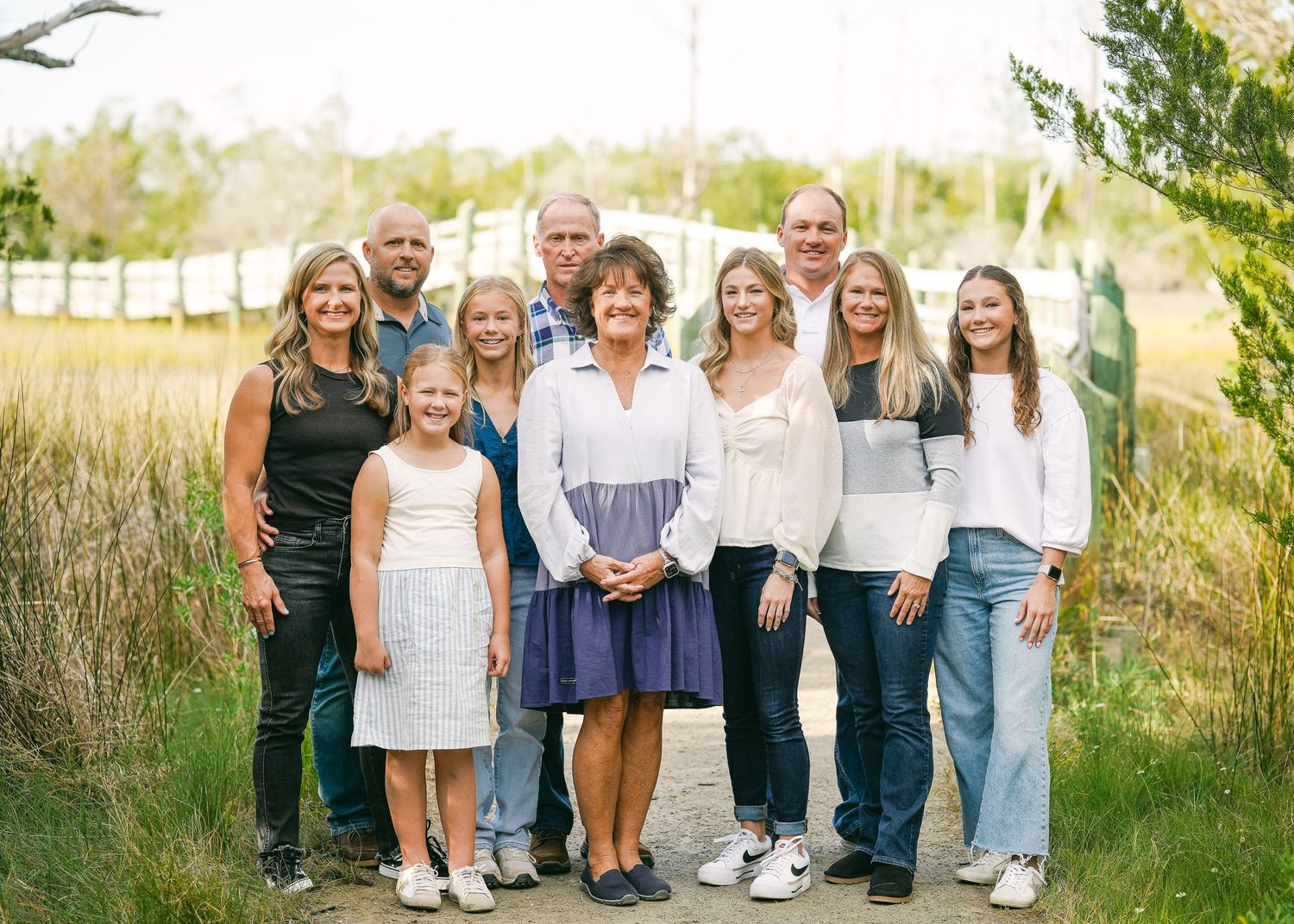Large family group portrait smiling outdoors in a lush green field near a white fence.