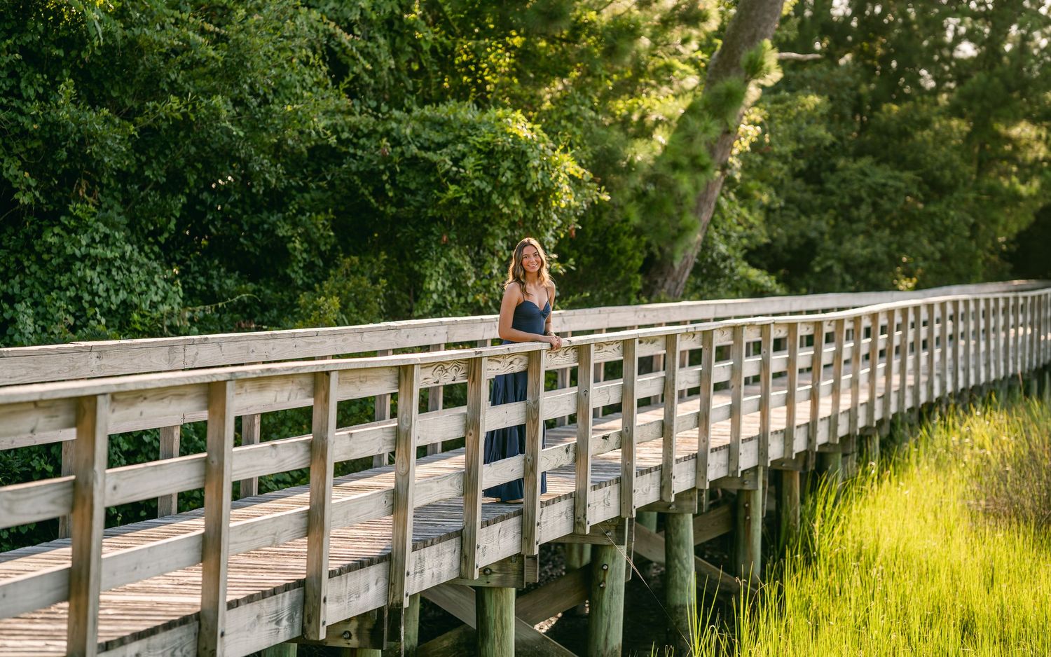 Woman in navy maxi dress standing on long wooden boardwalk surrounded by green marsh landscape.