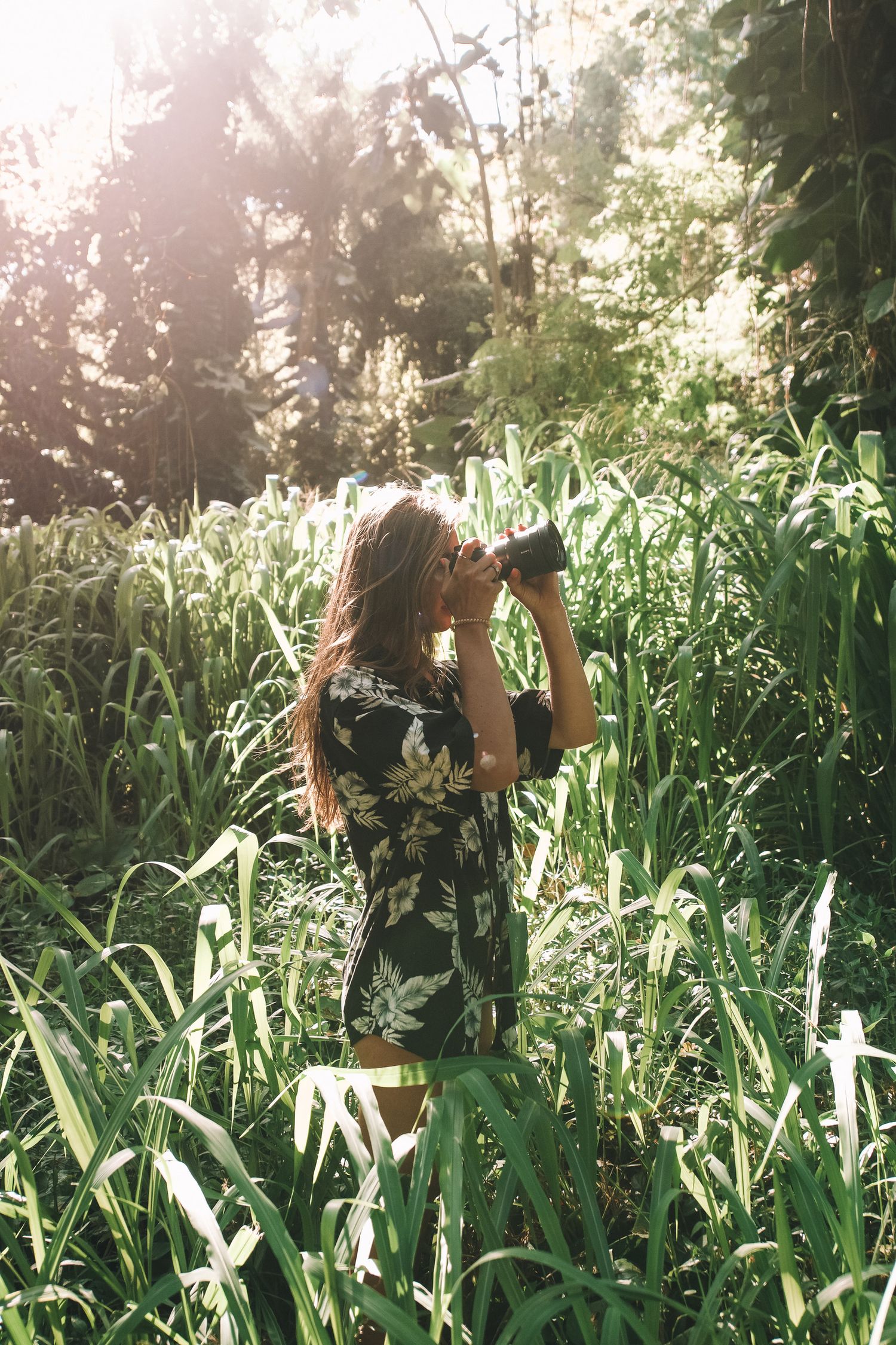 Young woman photographing with camera in lush green tropical forest with sunlight streaming through trees.