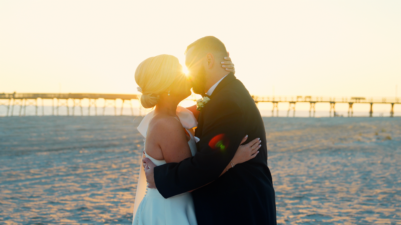 Couple sharing intimate kiss on beach at golden hour with pier in background.