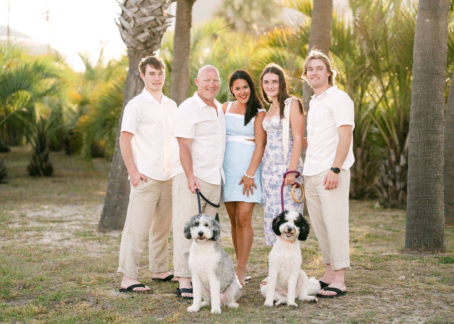 Smiling family of five standing with two fluffy black and white dogs among palm trees outdoors.
