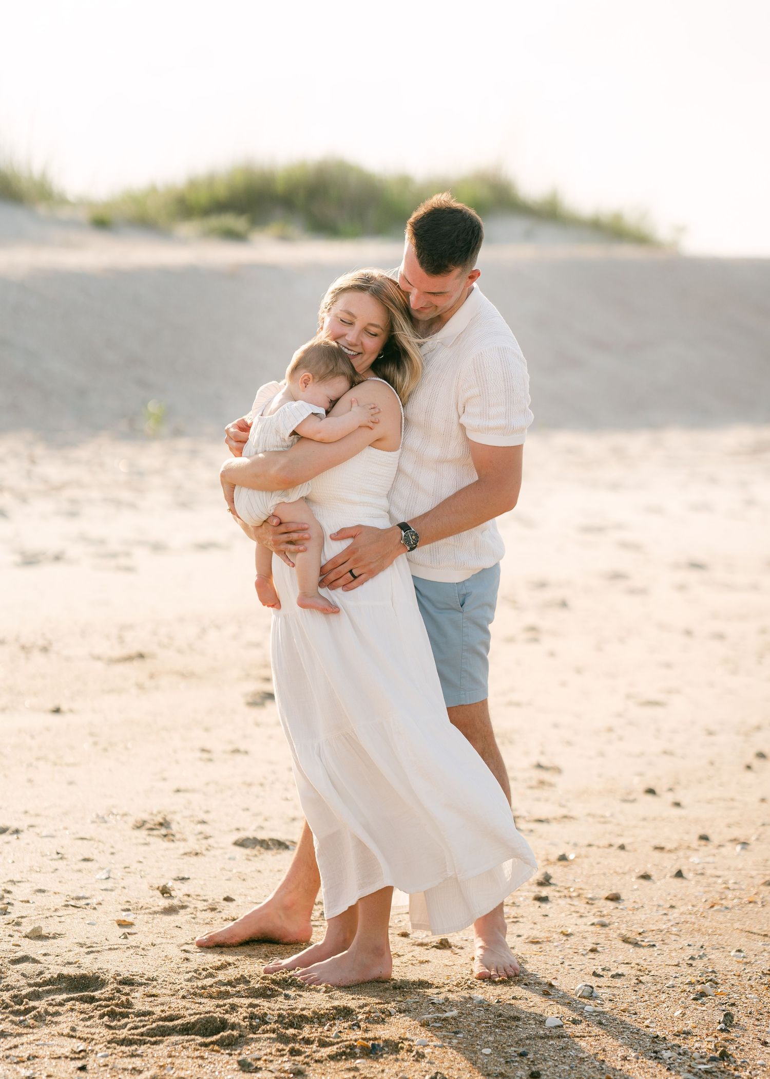 Loving family of three embracing on sandy beach, father in white shirt and blue shorts at sunset.