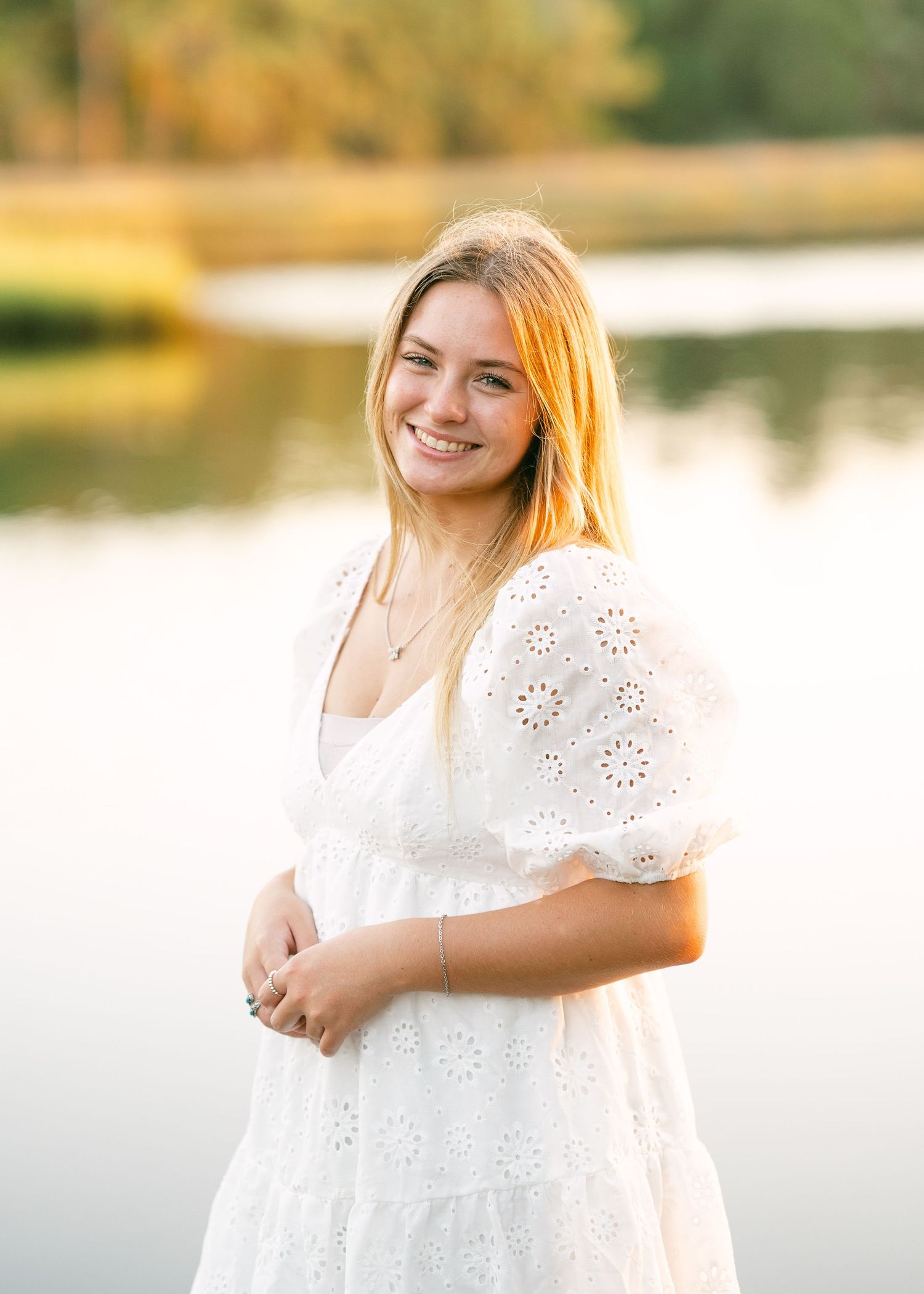 Blonde teen girl in white eyelet dress smiling by lake during golden hour senior photo.