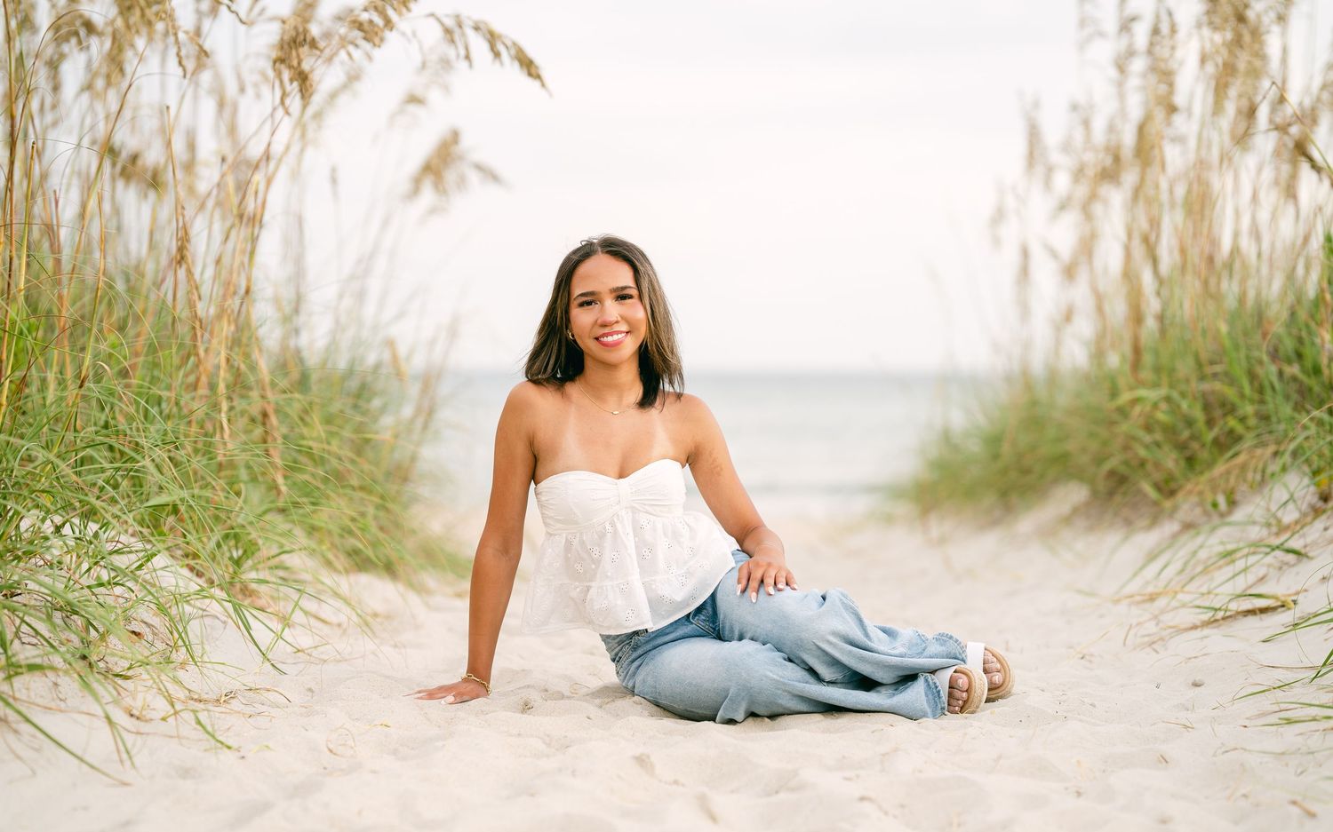 Smiling young woman in white strapless top and jeans sits cross-legged on sandy beach path with sea oats.
