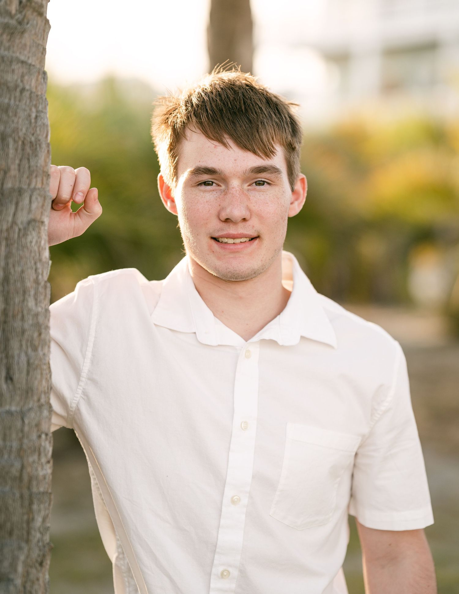 Smiling young man in white shirt leaning against a tree in a sunny outdoor tropical setting.