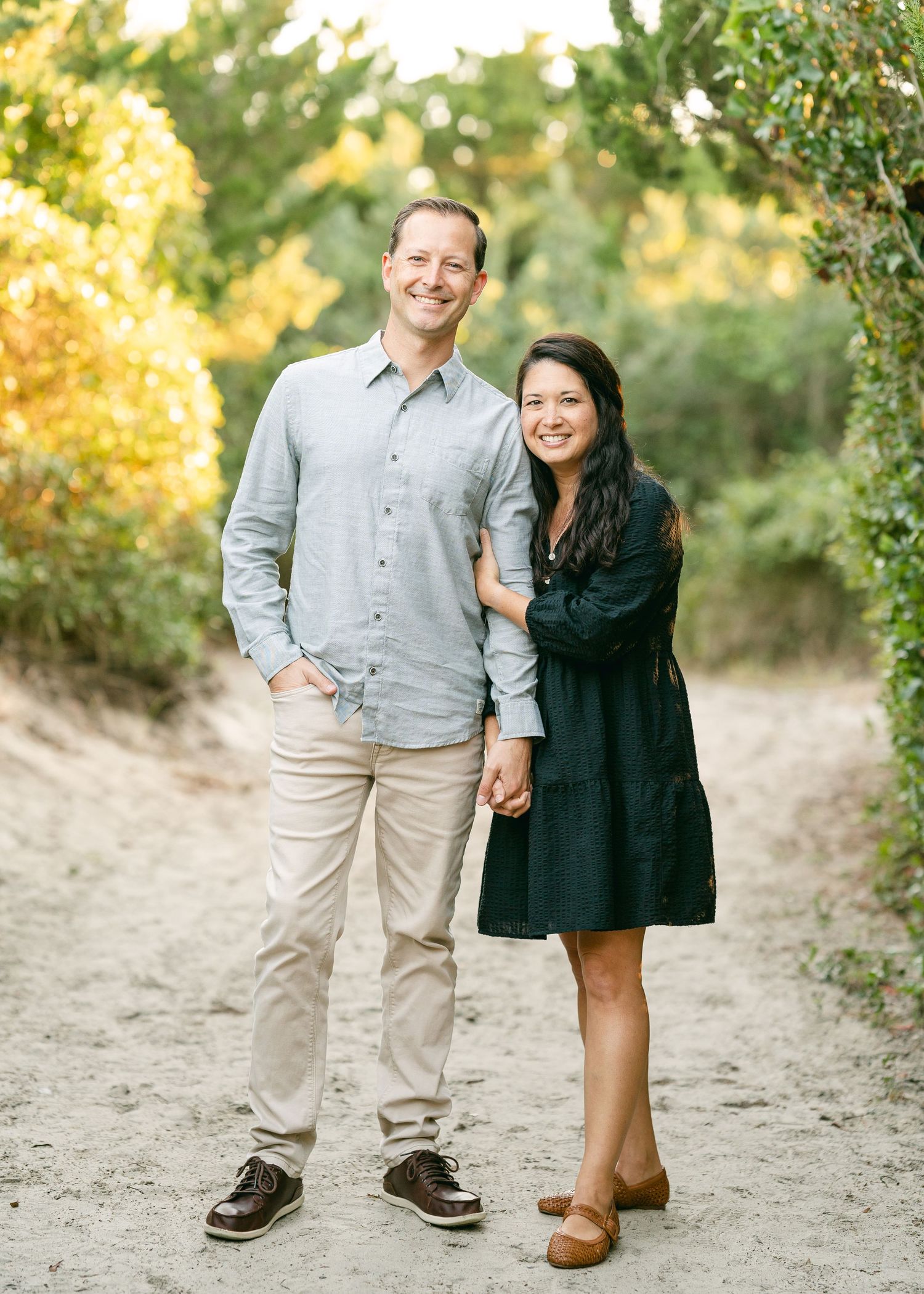 Happy couple holding hands and smiling on a sandy trail with golden foliage behind them.
