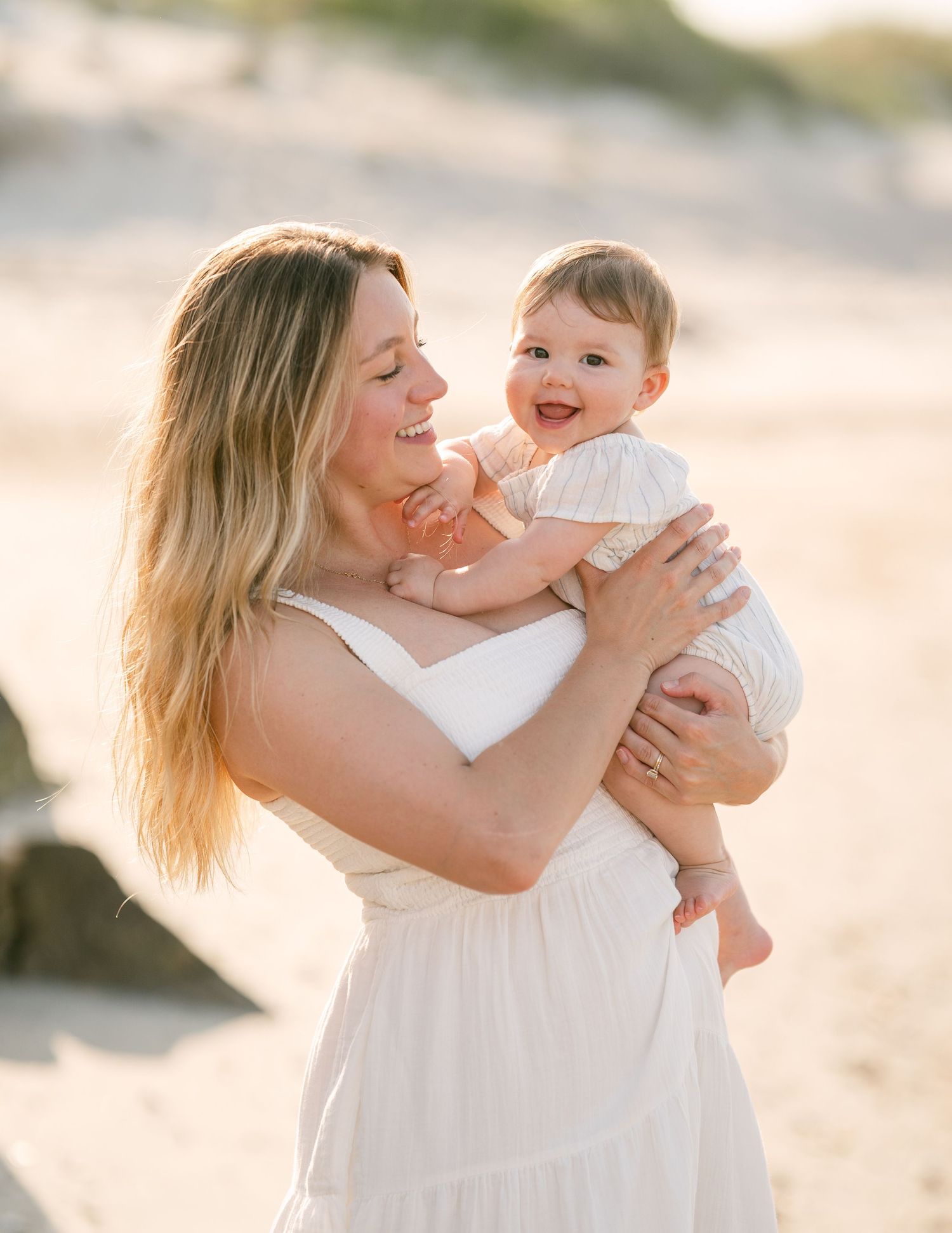 Blonde mother in white dress laughing with smiling baby on golden sandy beach.