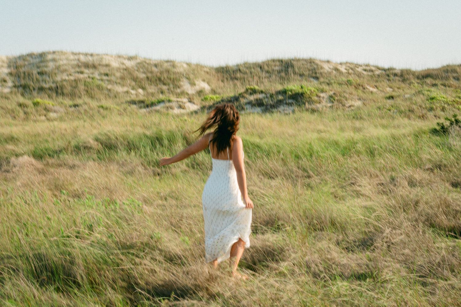 Woman in white dress running through windswept coastal grass and dunes.