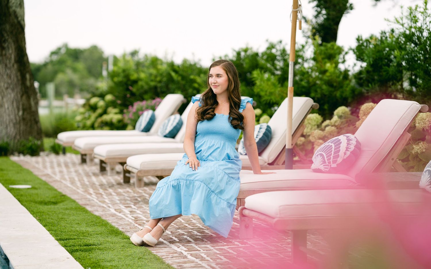 Woman in blue ruffle dress sitting on pink lounge chair by pool with lush greenery background.