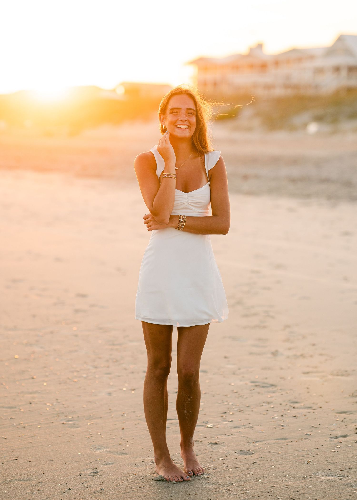 Smiling young woman in white mini dress standing barefoot on sandy beach at golden sunset.