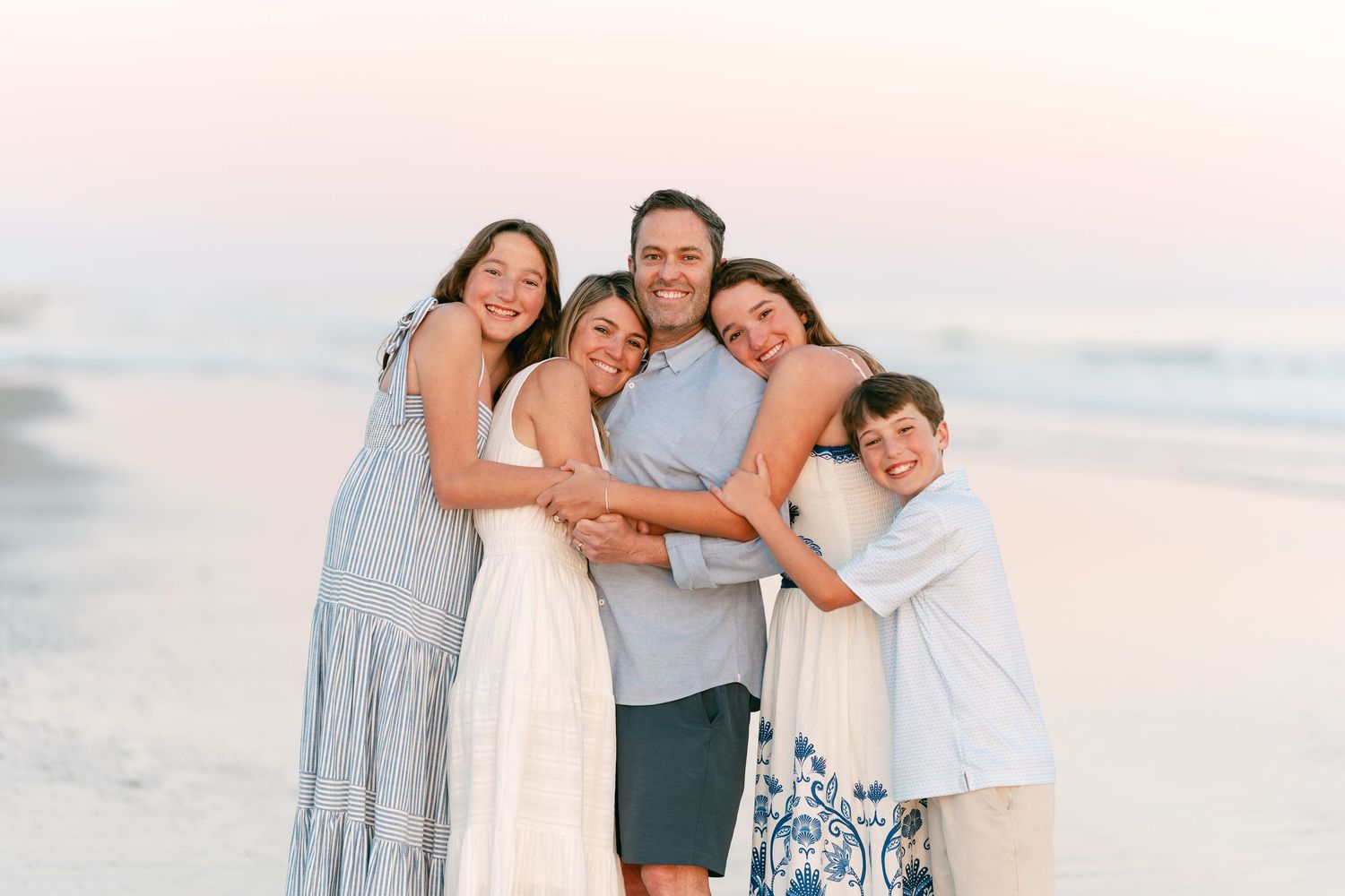 Joyful family of five hugging and laughing together on a sandy beach at sunset.