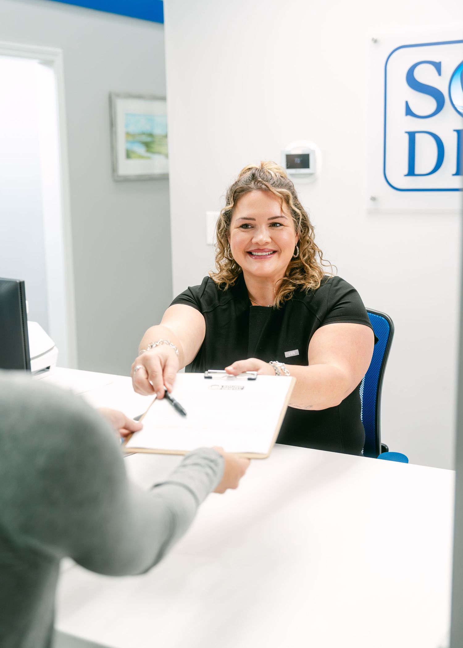 Smiling receptionist handing clipboard to patient at dental office front desk.