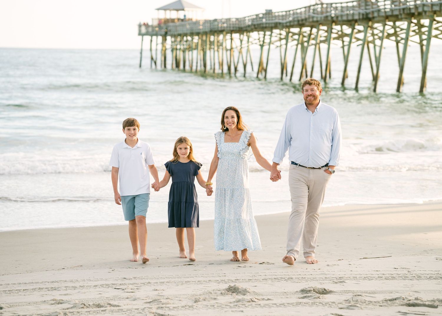 A family of four holds hands walking on a sandy beach near a wooden pier at sunset.