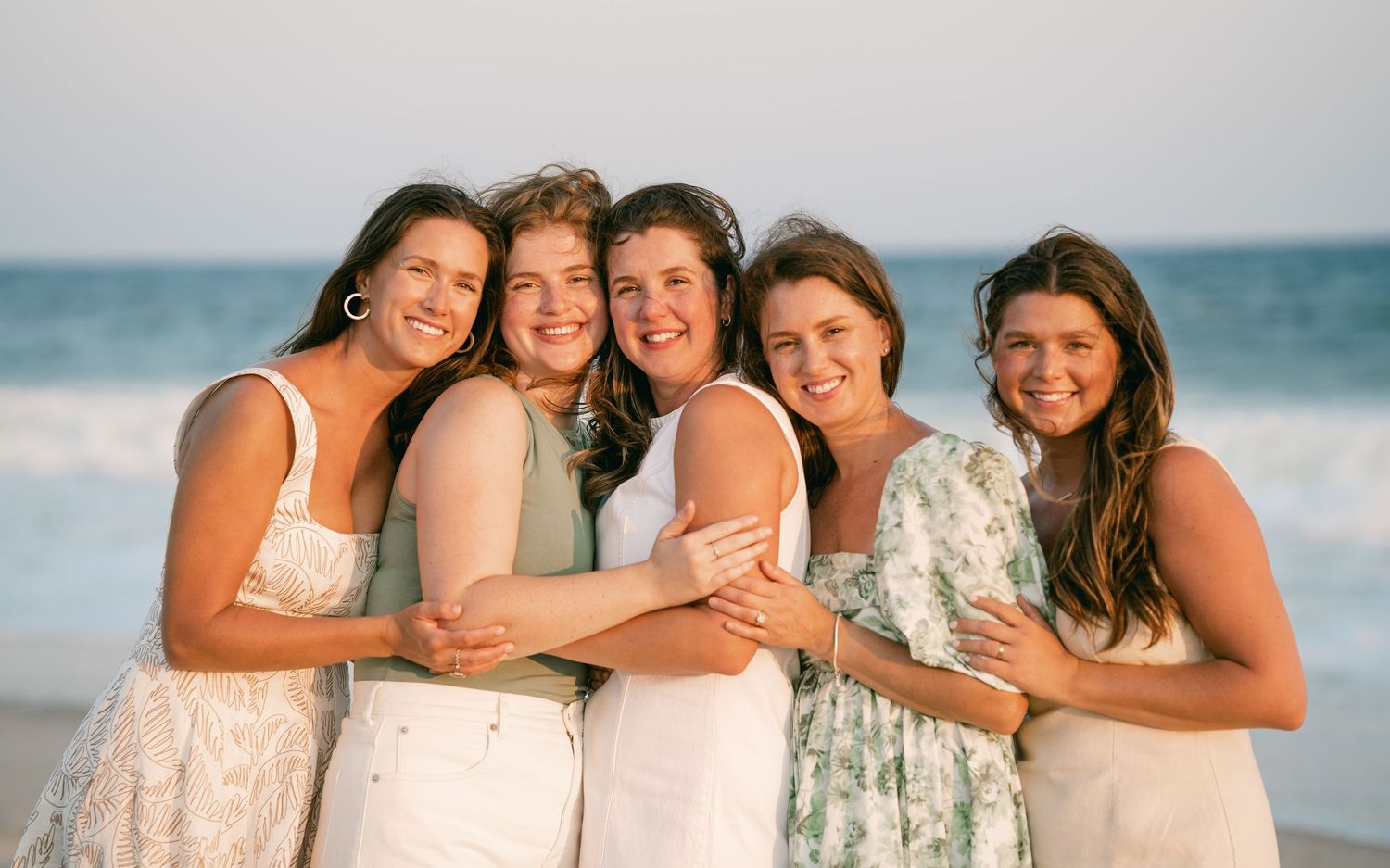 Five women smiling and embracing each other on a beach at sunset.