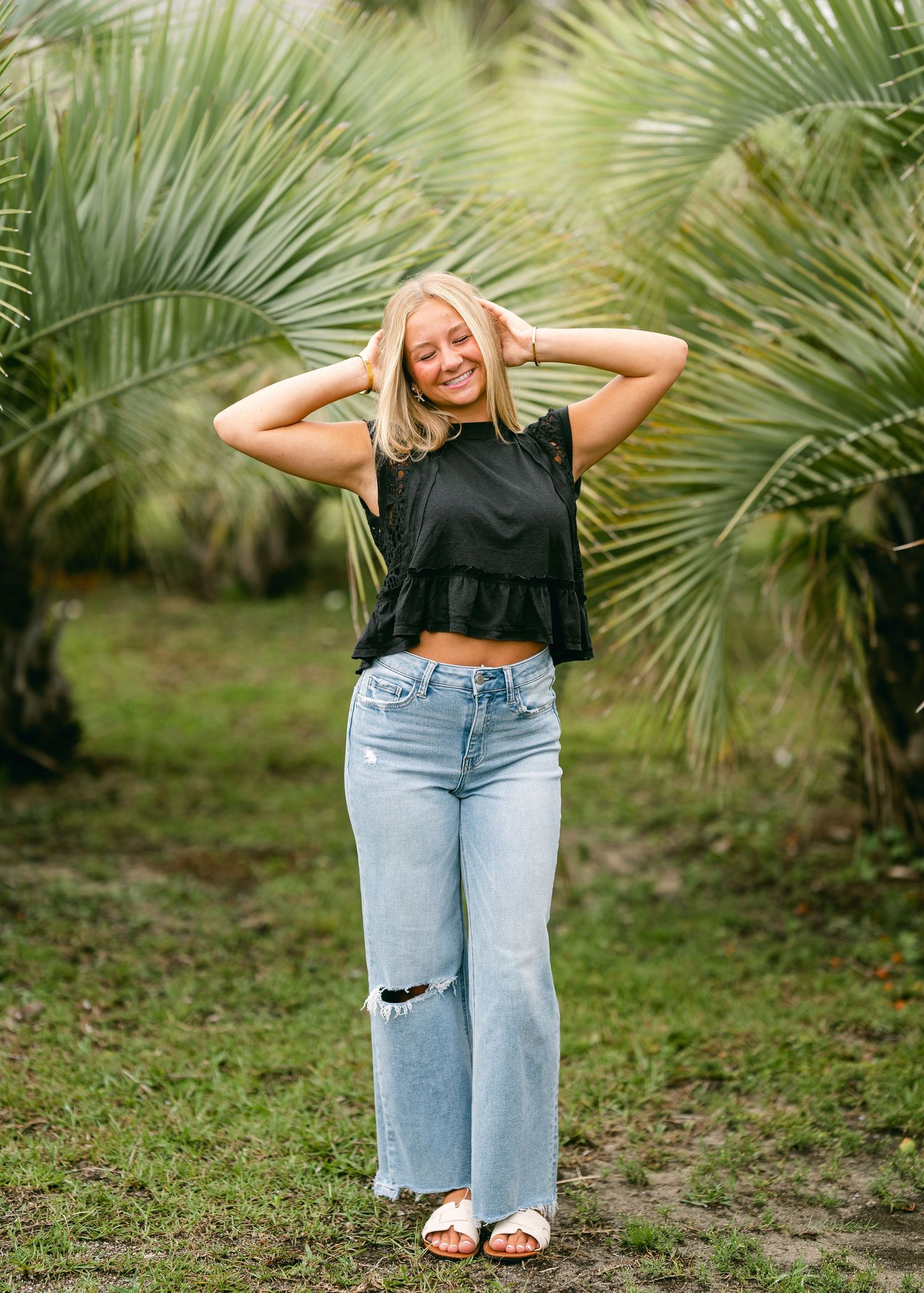 Laughing blonde teen girl in black crop top and ripped wide-leg jeans poses among lush tropical palm trees.