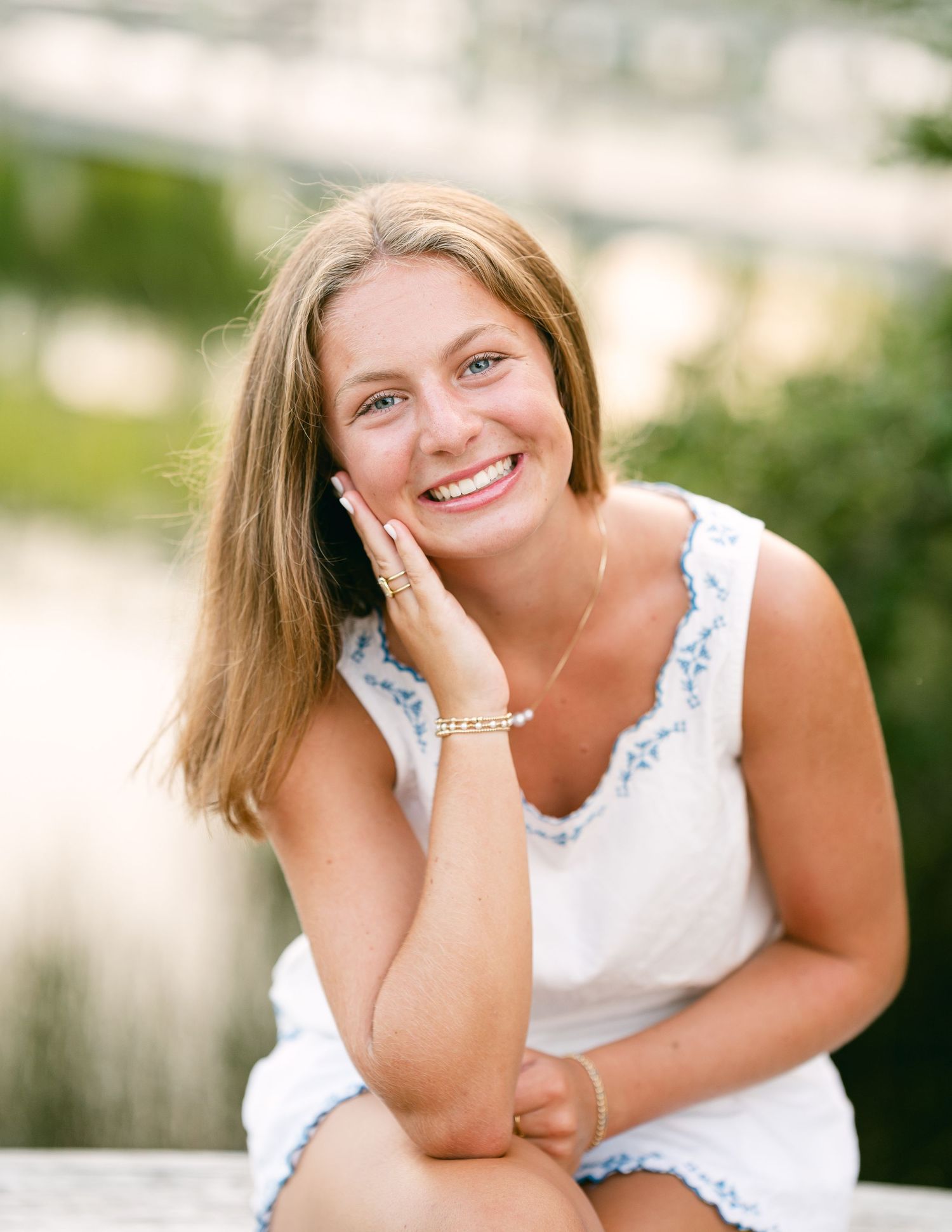 Smiling teenage girl in white dress posing outdoors near water for senior portrait.