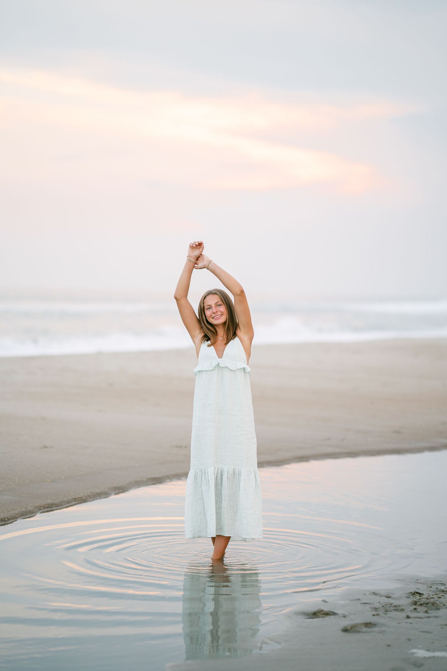 Woman in white maxi dress standing in shallow beach water with arms raised at sunset.