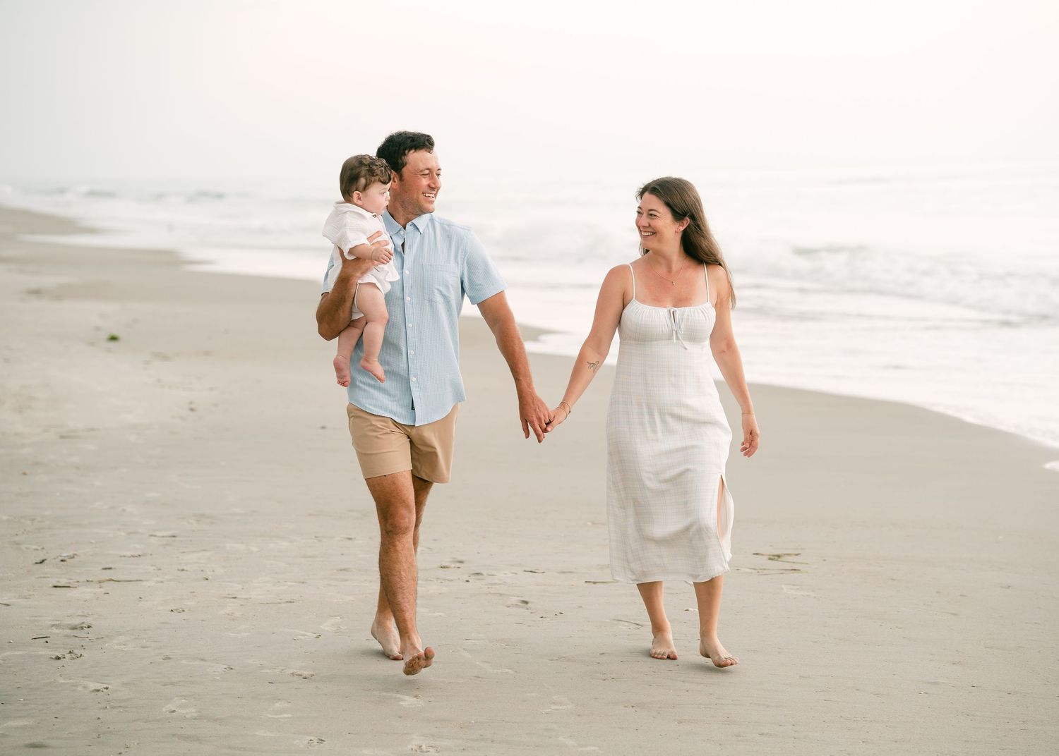 Young couple holding hands walking on beach while man carries infant at golden hour.