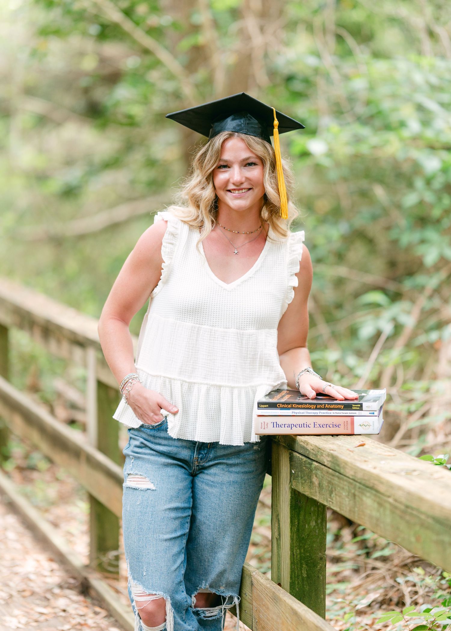 Blonde graduate in white top and jeans wearing cap holding textbooks on wooden bridge.