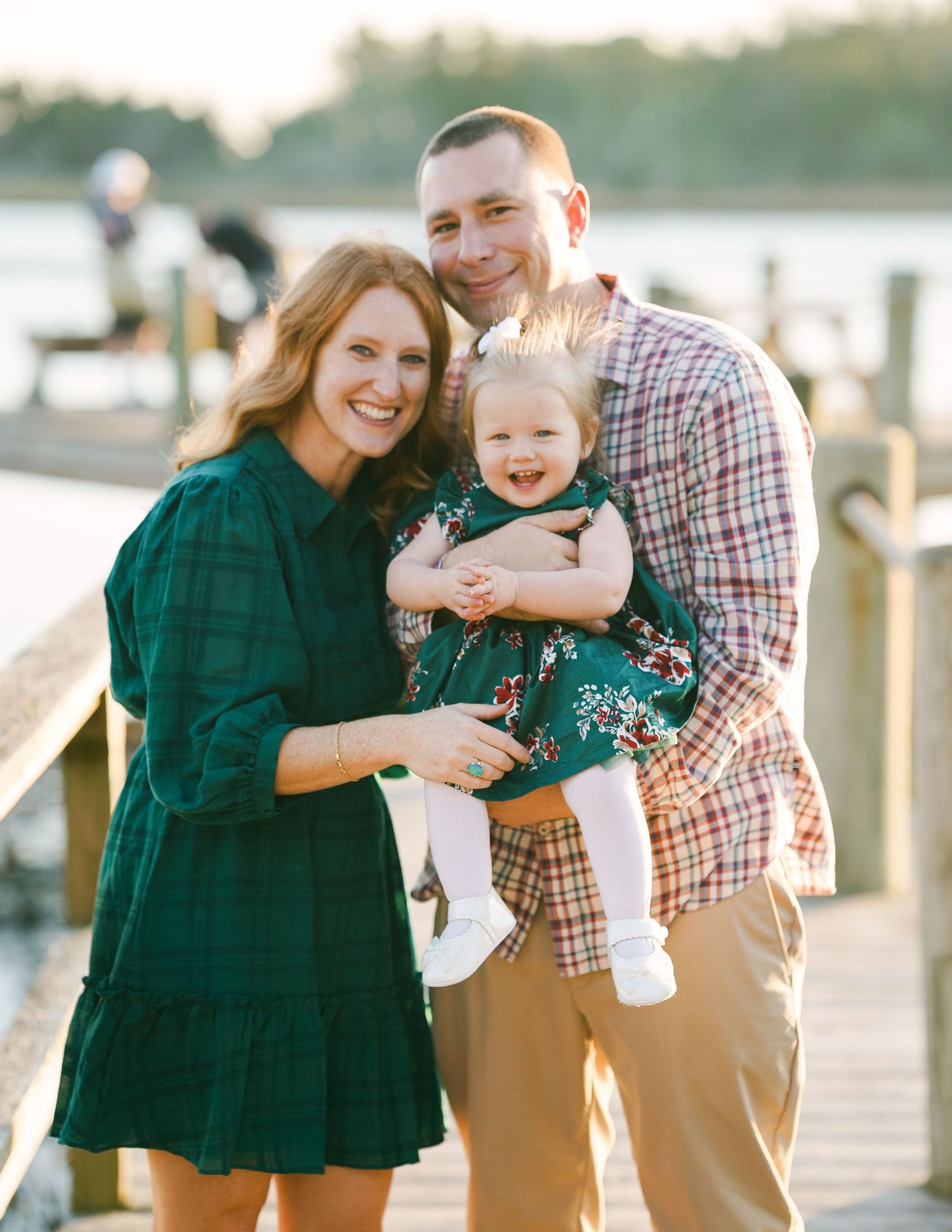 Happy young family with parents holding laughing baby girl on a waterfront dock at sunset.