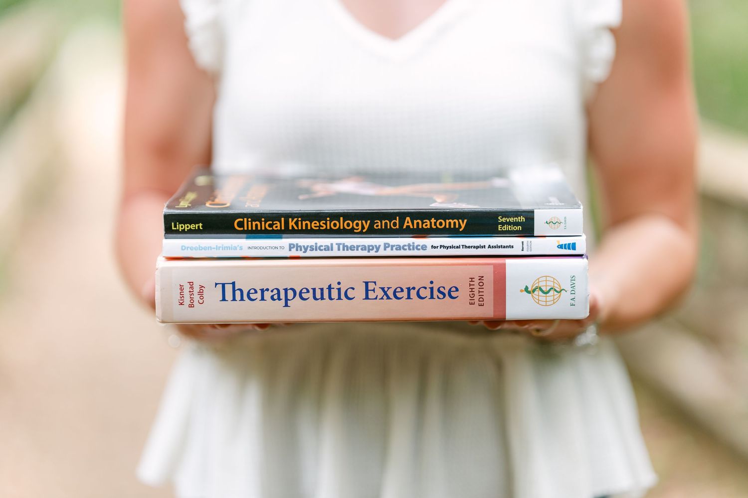 Student holds physical therapy textbooks including Therapeutic Exercise and Clinical Kinesiology outdoors.