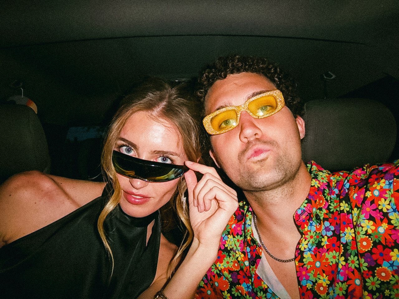 Couple wearing colorful sunglasses taking a selfie in a car, man in floral shirt and woman in black top.