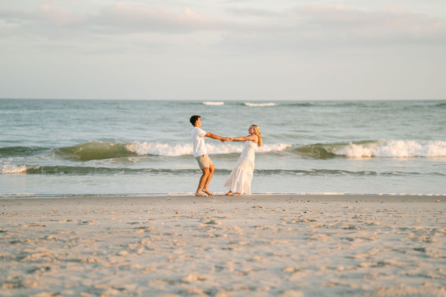 Couple dancing and spinning together on sandy beach with ocean waves at golden hour.