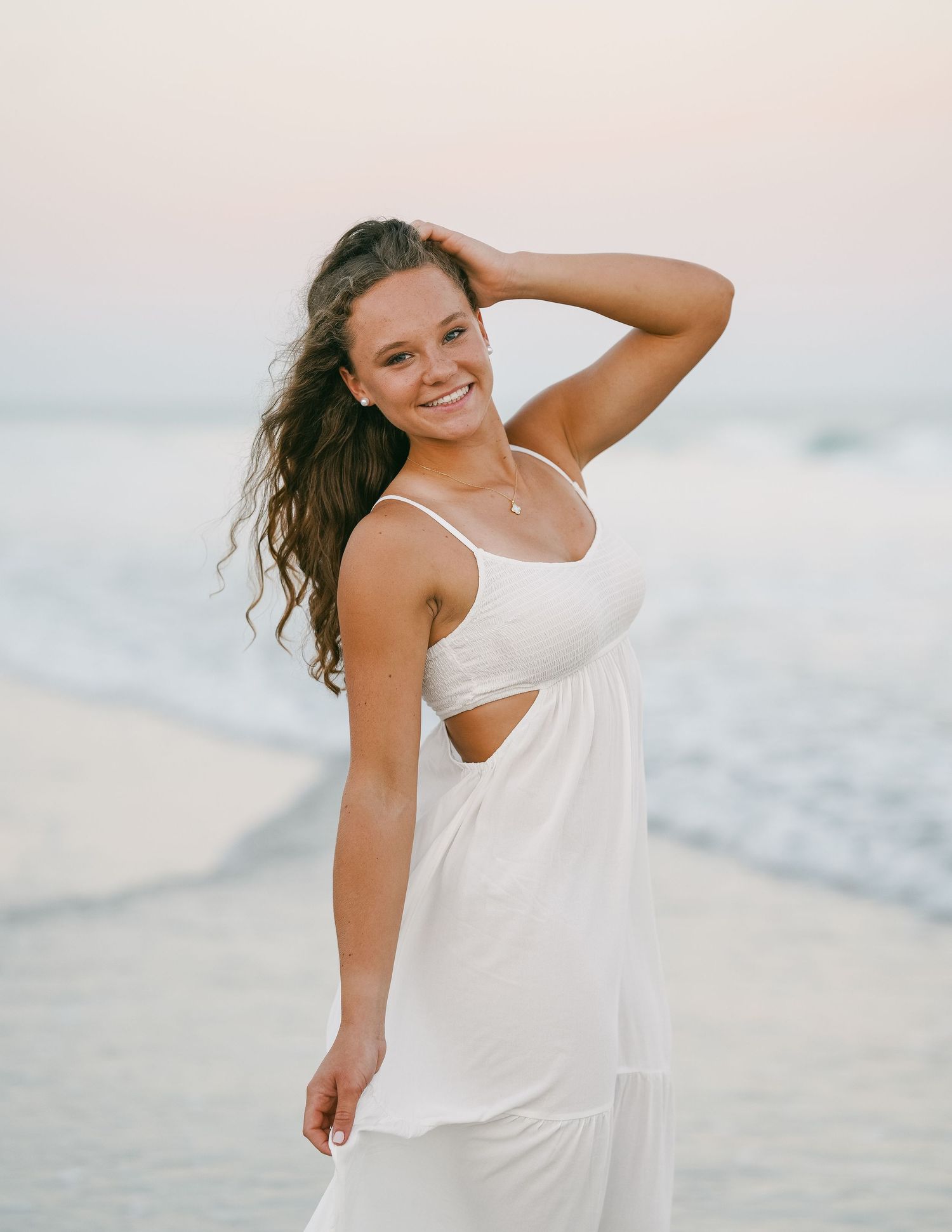 Smiling teen girl in white cutout dress posing on beach at sunset for senior portrait.