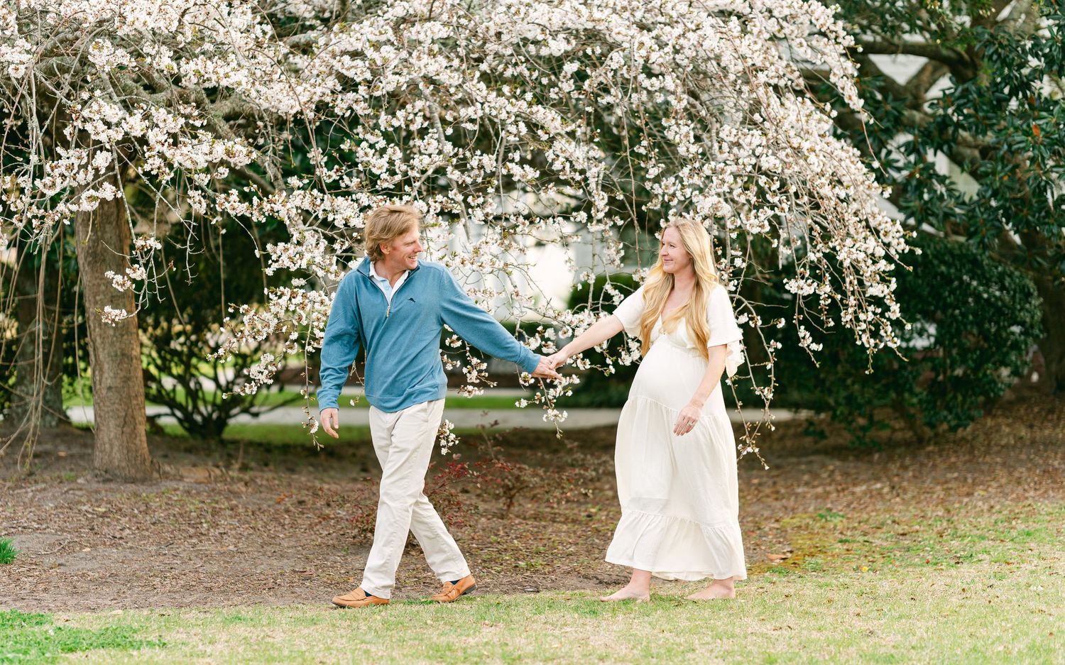 Couple walking hand in hand under blooming white cherry blossom trees in a garden.